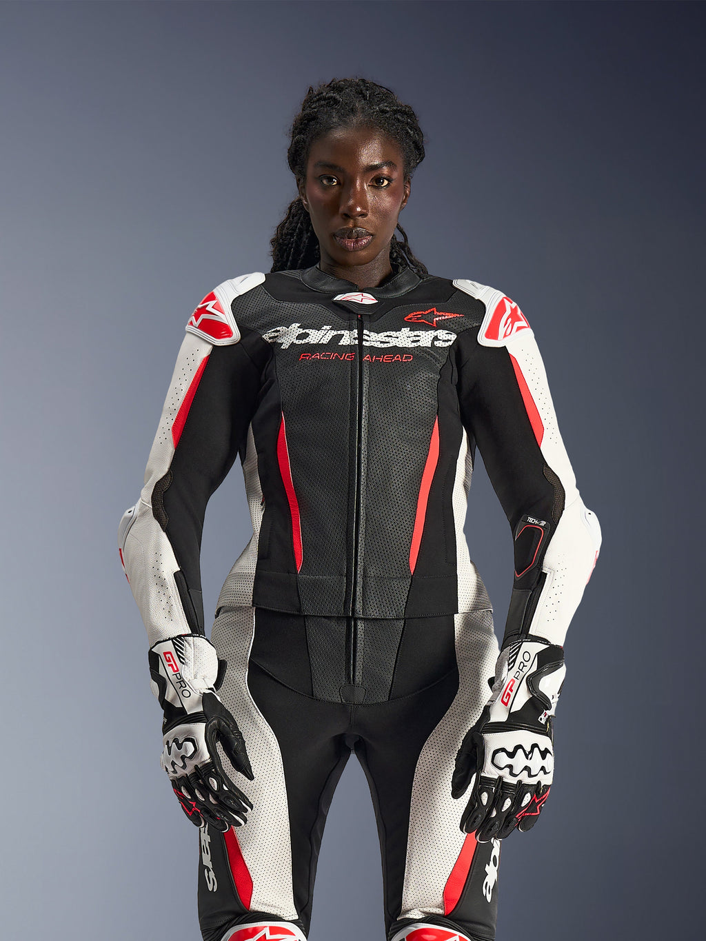 Kvinnlig modell i Alpinestars Stella GP-R7 2-delat läderställ i svart, vitt och rött fluo, med perforerade läderpaneler och matchande GP Pro-handskar, anatomiskt optimerat för kvinnliga förare.
