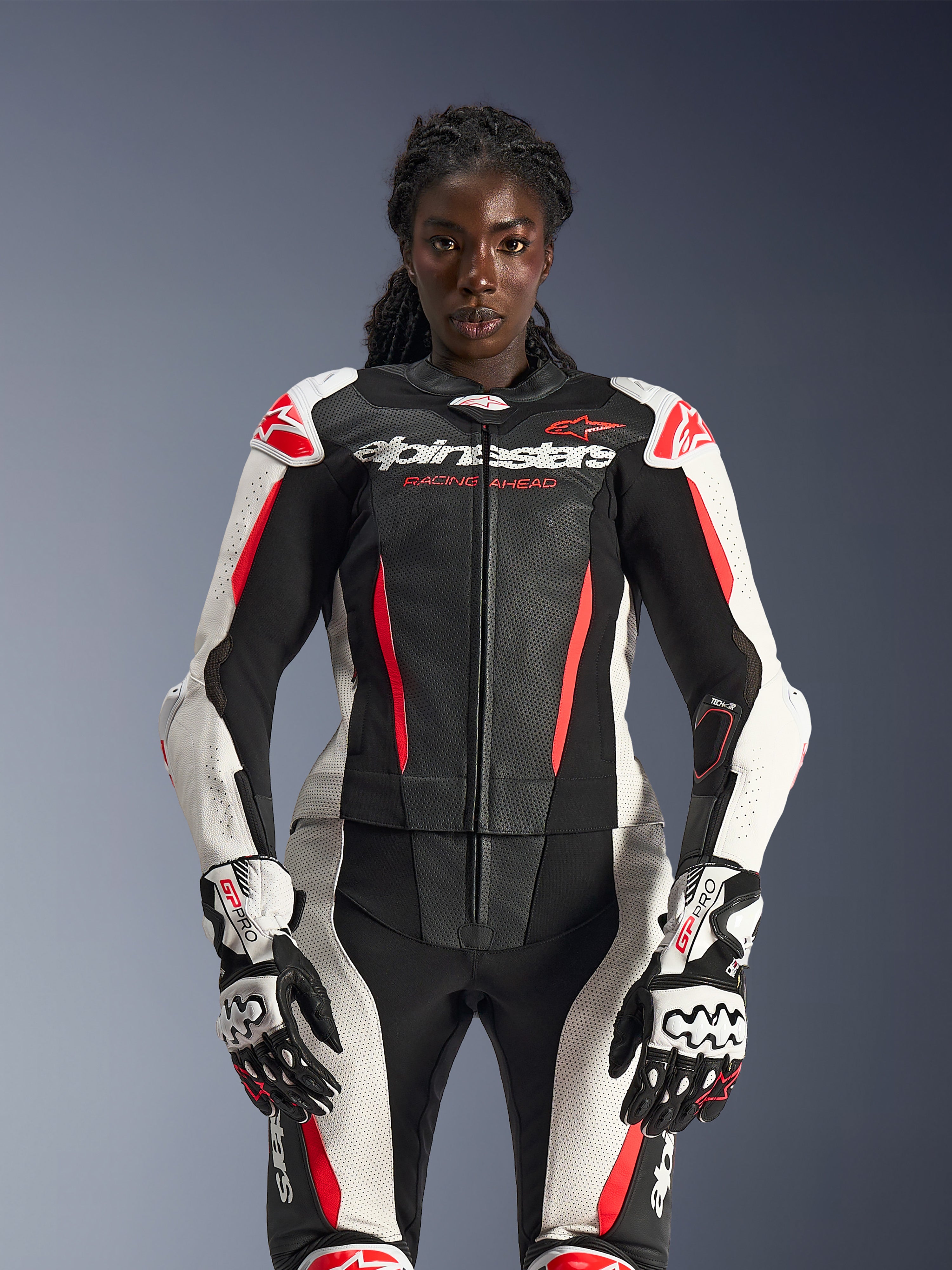 Kvinnlig modell i Alpinestars Stella GP-R7 2-delat läderställ i svart, vitt och rött fluo, med perforerade läderpaneler och matchande GP Pro-handskar, anatomiskt optimerat för kvinnliga förare.