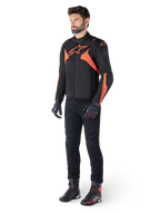 Alpinestars T-Jaws V4 Waterproof Jacket, svart och röd fluo, bärs av en manlig modell med svarta byxor, handskar och stövlar, med lysande orangea detaljer och Alpinestars-logotyp, sportig design för motorcykelkörning