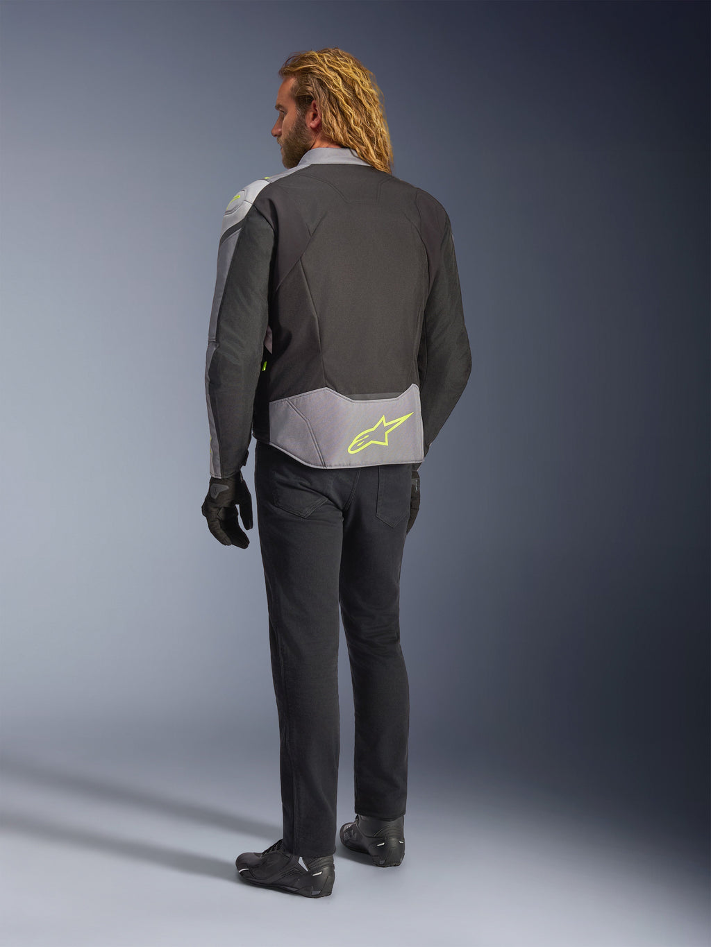 Alpinestars T-Jaws V4 Waterproof Jacket, back view, Black Ice Gray and Dark Gray, featuring a neon yellow Alpinestars logo on the lower back, worn with black pants and motorcycle gloves, sport-styled riding apparel