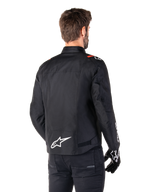 Alpinestars T-SPS V2 Waterproof-jacka, Black White Red Fluo, sedd bakifrån, med vit Alpinestars-logotyp på nedre delen av ryggen och röda detaljer på axlarna, bärs med svarta körhandskar och byxor, sportig textiljacka för motorcykel.