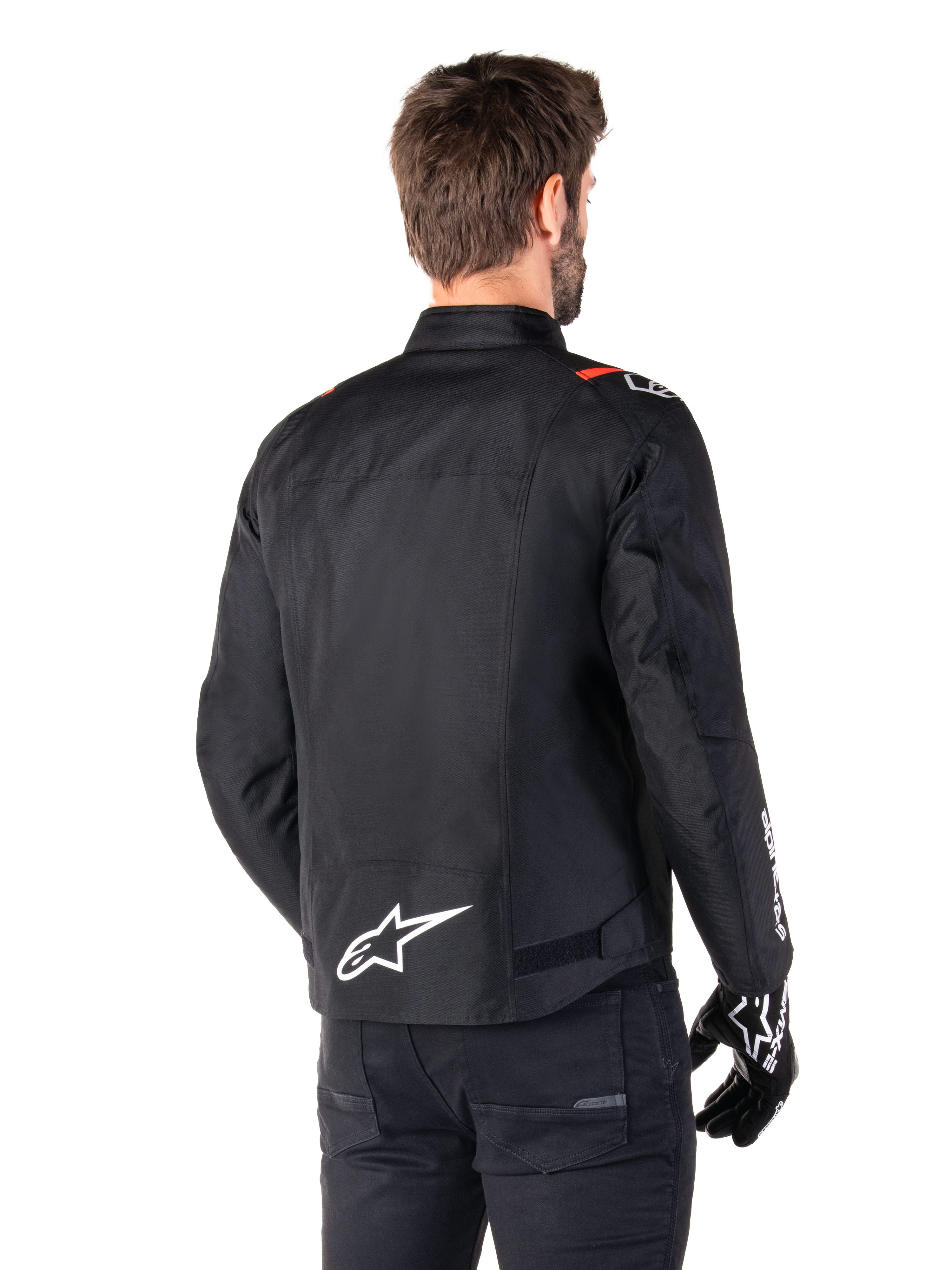 Alpinestars T-SPS V2 Waterproof-jacka, Black White Red Fluo, sedd bakifrån, med vit Alpinestars-logotyp på nedre delen av ryggen och röda detaljer på axlarna, bärs med svarta körhandskar och byxor, sportig textiljacka för motorcykel.