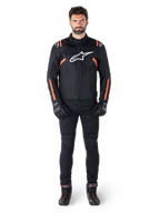 Manlig modell som bär Alpinestars T-SPS V2 Waterproof-jacka i Black White Red Fluo, kombinerad med svarta motorcykelbyxor och handskar, med en vit Alpinestars-logotyp på bröstet och röda detaljer på ärmarna.