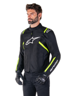 Manlig modell bär Alpinestars T-SPS V2 Waterproof Jacket, svart med vita och neongula detaljer, vit Astars-logotyp på bröstet och fluorescerande gula ränder på axlar och ärmar, svarta byxor och körhandskar, sportig textiljacka för motorcykel.