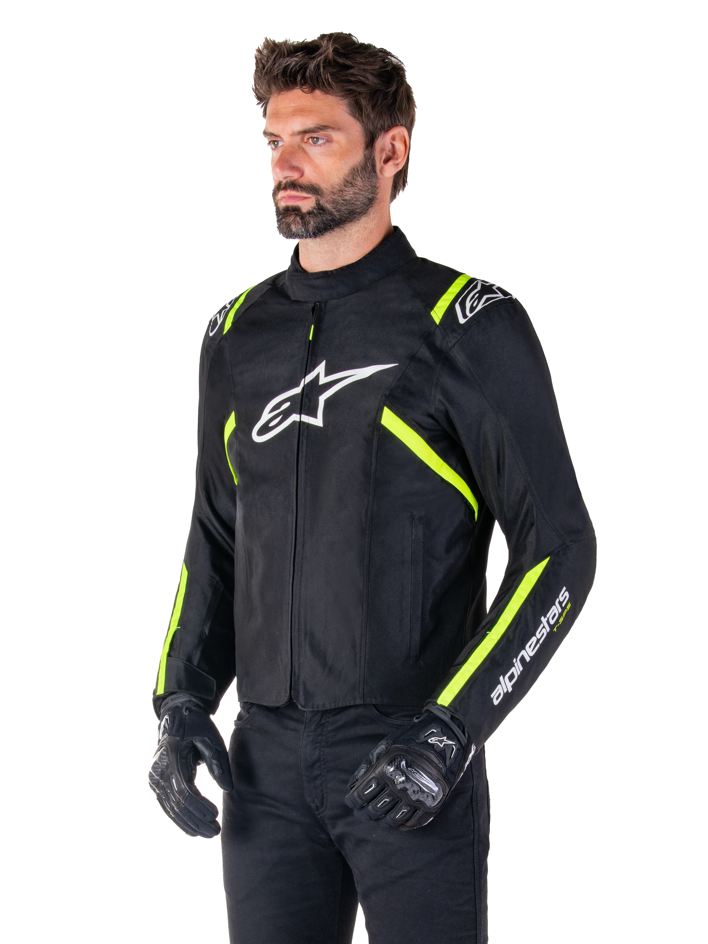 Manlig modell bär Alpinestars T-SPS V2 Waterproof Jacket, svart med vita och neongula detaljer, vit Astars-logotyp på bröstet och fluorescerande gula ränder på axlar och ärmar, svarta byxor och körhandskar, sportig textiljacka för motorcykel.