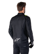 Alpinestars T-SPS V2 Waterproof Jacket, svart vit gul fluo, bakifrån vy av manlig modell som bär motorcykeljackan i textil, med vit logotyp på nedre delen av ryggen och neongula detaljer på axlarna, kombinerat med svarta byxor och handskar.