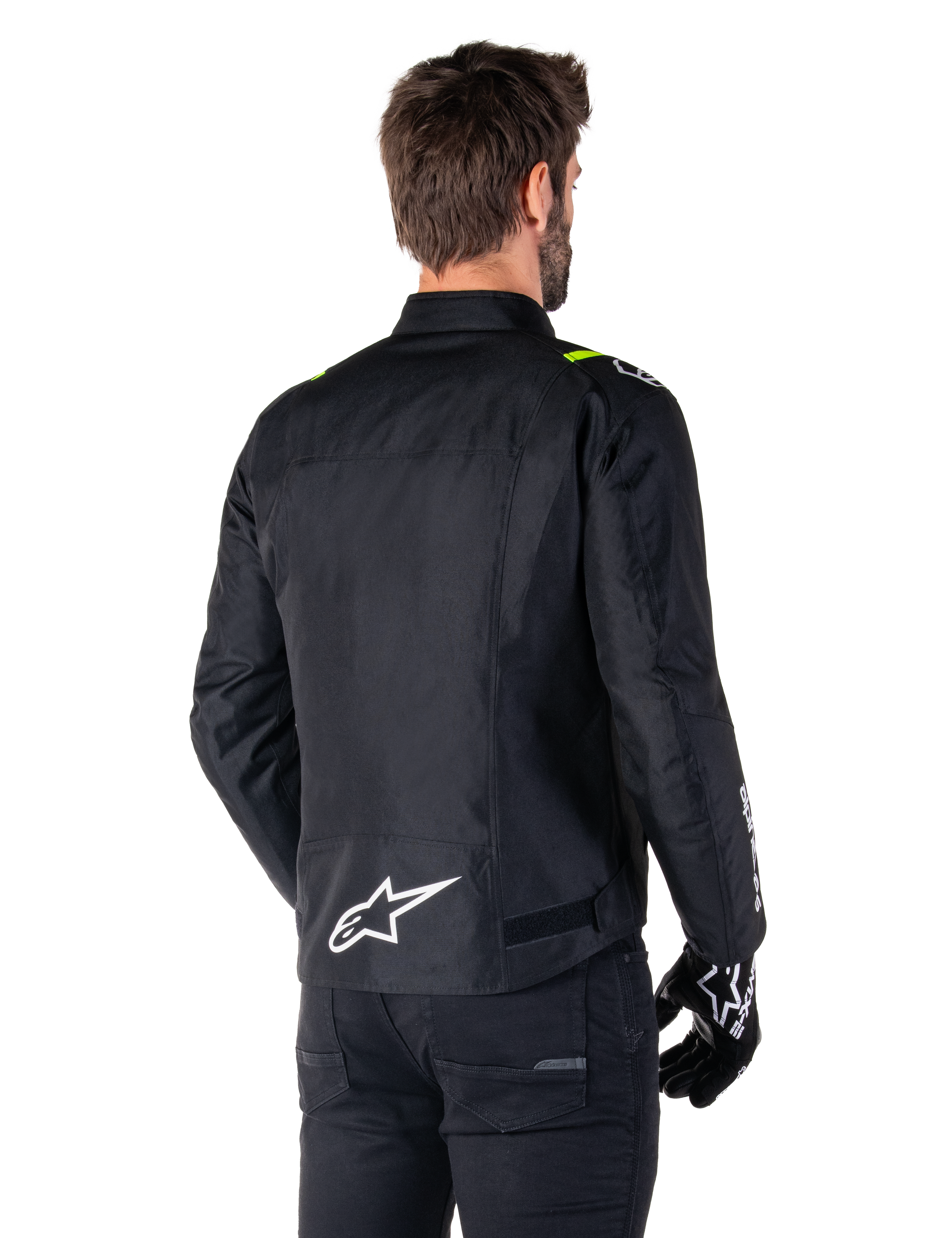 Alpinestars T-SPS V2 Waterproof Jacket, svart vit gul fluo, bakifrån vy av manlig modell som bär motorcykeljackan i textil, med vit logotyp på nedre delen av ryggen och neongula detaljer på axlarna, kombinerat med svarta byxor och handskar.