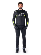 Alpinestars T-SPS V2 Vattentät Jacka, svart, vit och gul fluo, bärs av en modell med svarta körbyxor och handskar, vit Alpinestars-logotyp på bröstet och fluorescerande detaljer på axlar och ärmar, sportig motorcykelutrustning