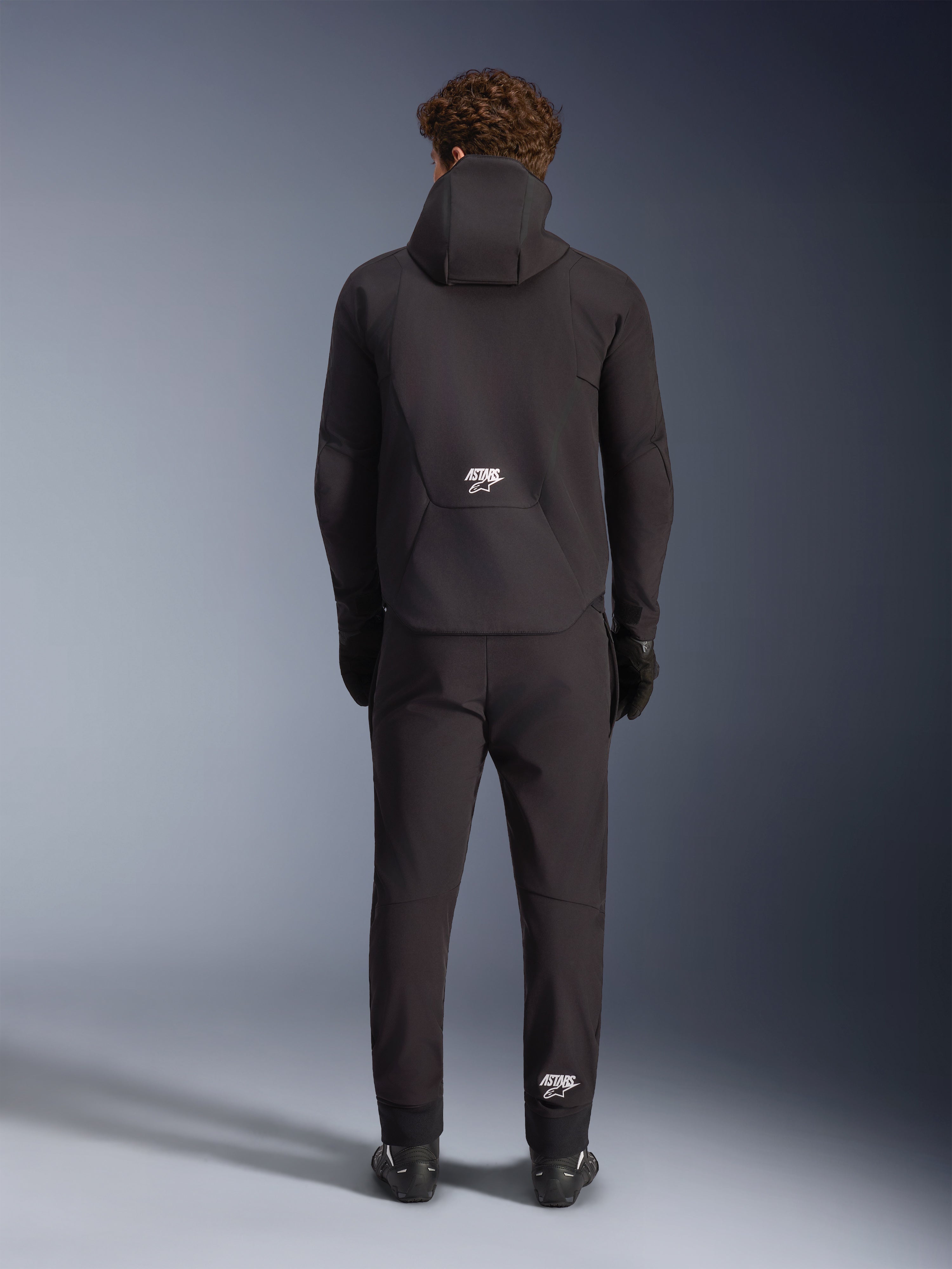 Alpinestars Aeroshell Waterproof Jacket, Black, seen from the back on a model wearing matching black riding pants and gloves, hooded sports softshell design with white ASTARS logos, sleek and modern motorcycle apparel