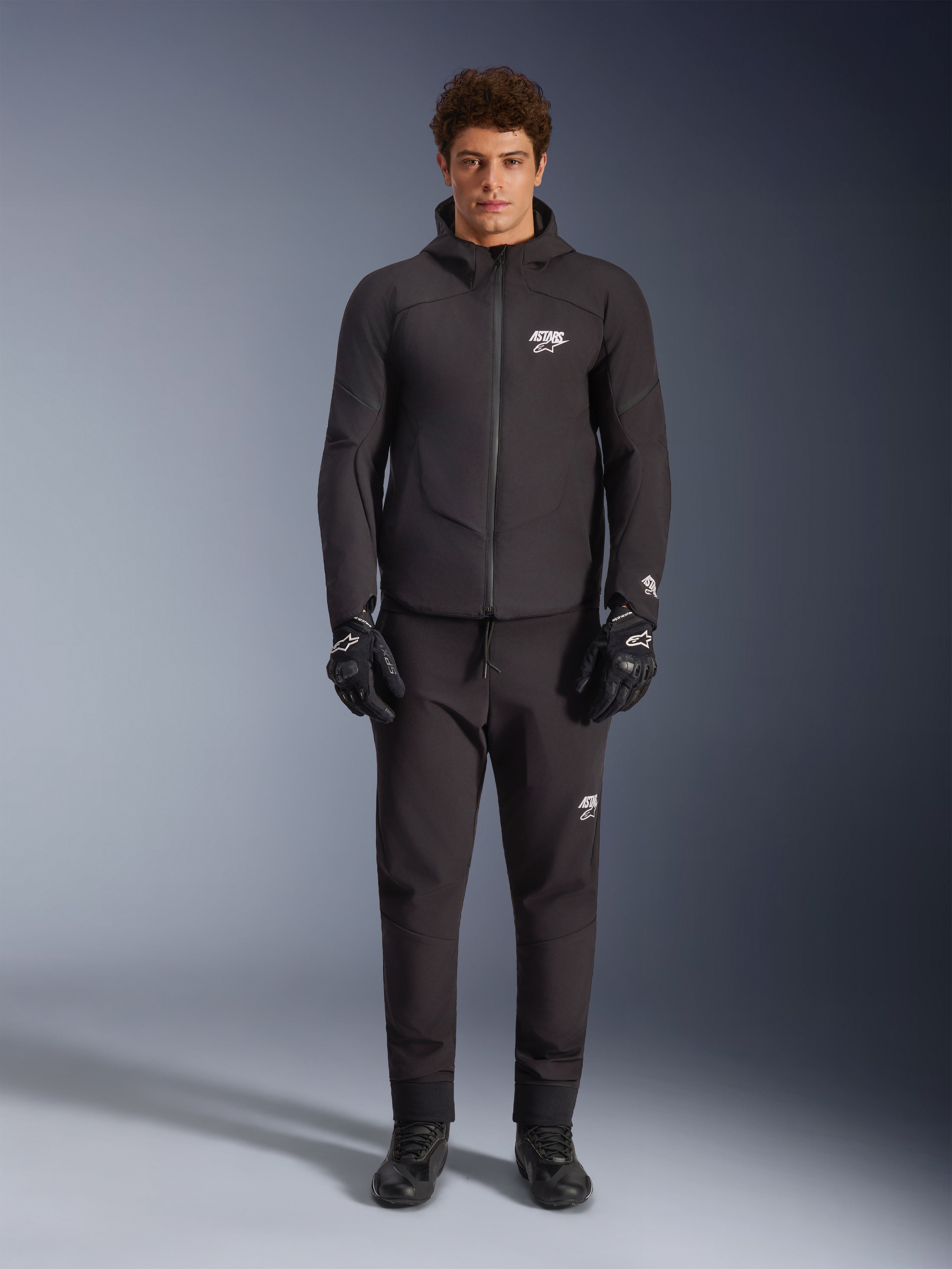 Male model wearing Alpinestars Aeroshell Waterproof Jacket in Black, paired with matching black technical pants and riding gloves, featuring a hooded softshell design with white logo accents, modern motorsport-inspired outfit for road riding