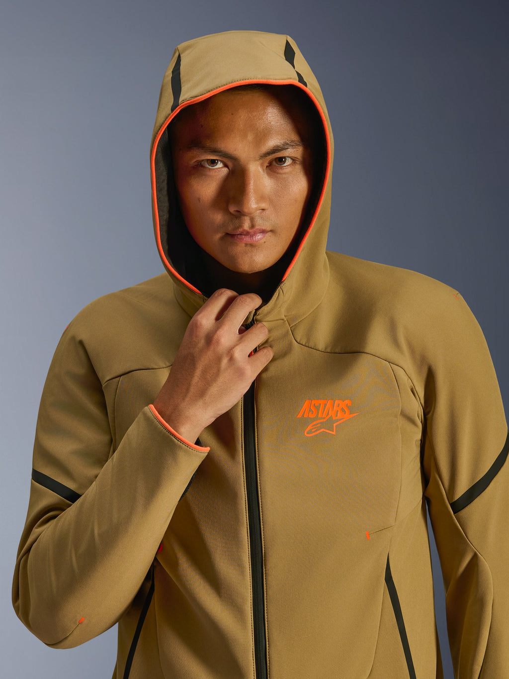 Alpinestars Aeroshell Waterproof Jacket, Dark Khaki Black Orange Fluo, model wearing the jacket with the hood up, featuring vibrant orange fluo trim and an orange ASTARS logo, sleek black accents, modern sport-styled waterproof softshell