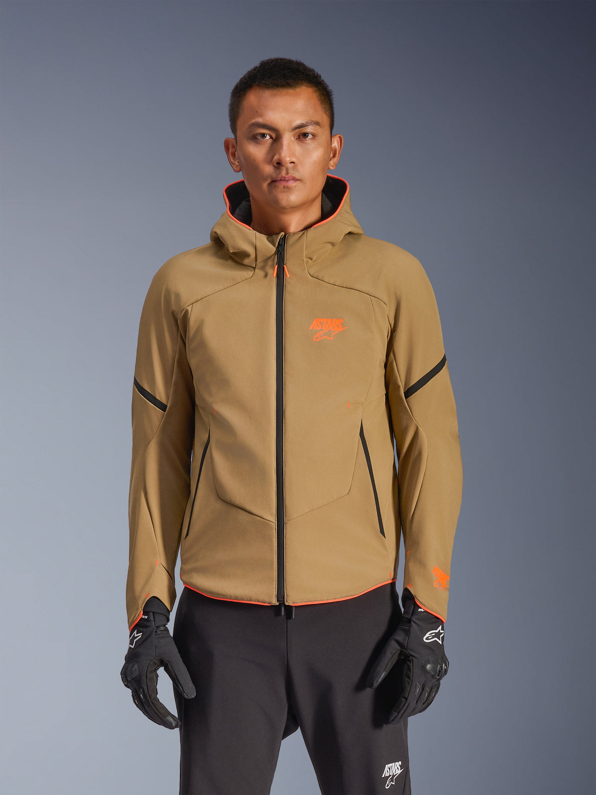 Alpinestars Aeroshell Waterproof Jacket, mörkhaki motorcykeljacka med huva, orange logotyper och svarta dragkedjor, svarta accentlinjer på ärmarna, visas med svarta handskar och byxor