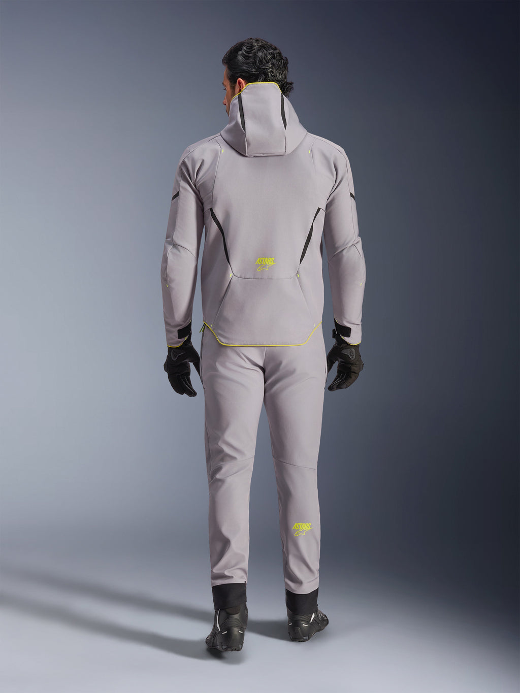 Alpinestars Aeroshell Waterproof Jacket i Mid Gray Yellow Fluo, vy bakifrån, bärs med matchande grå byxor, svarta handskar och svarta mc-skor. Har en softshelldesign med huva, fluorgula 'ASTARS'-detaljer och logotyper, konstruerad för modern motorcykelprestanda på väg.