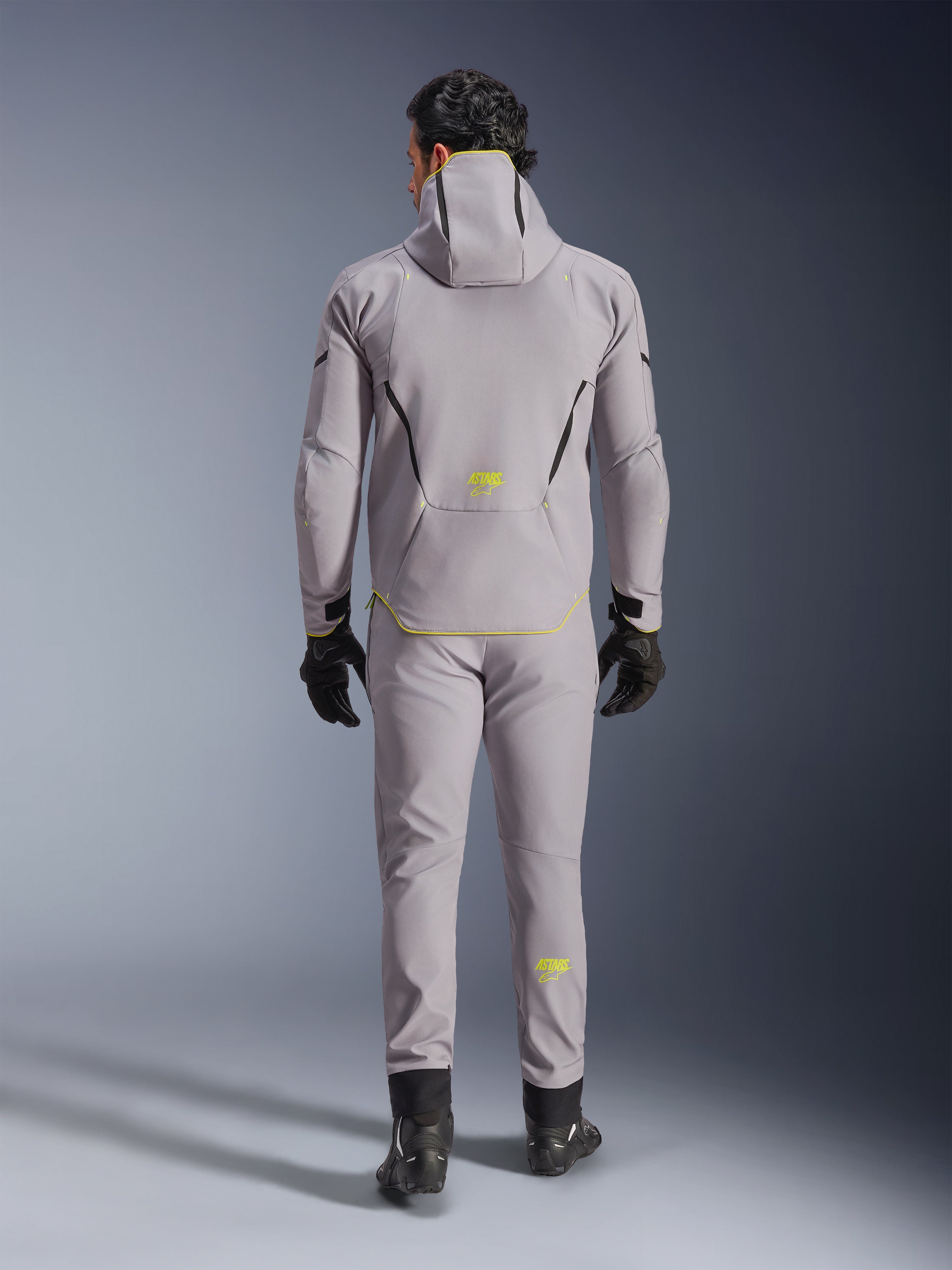 Alpinestars Aeroshell Waterproof Jacket i Mid Gray Yellow Fluo, vy bakifrån, bärs med matchande grå byxor, svarta handskar och svarta mc-skor. Har en softshelldesign med huva, fluorgula 'ASTARS'-detaljer och logotyper, konstruerad för modern motorcykelprestanda på väg.