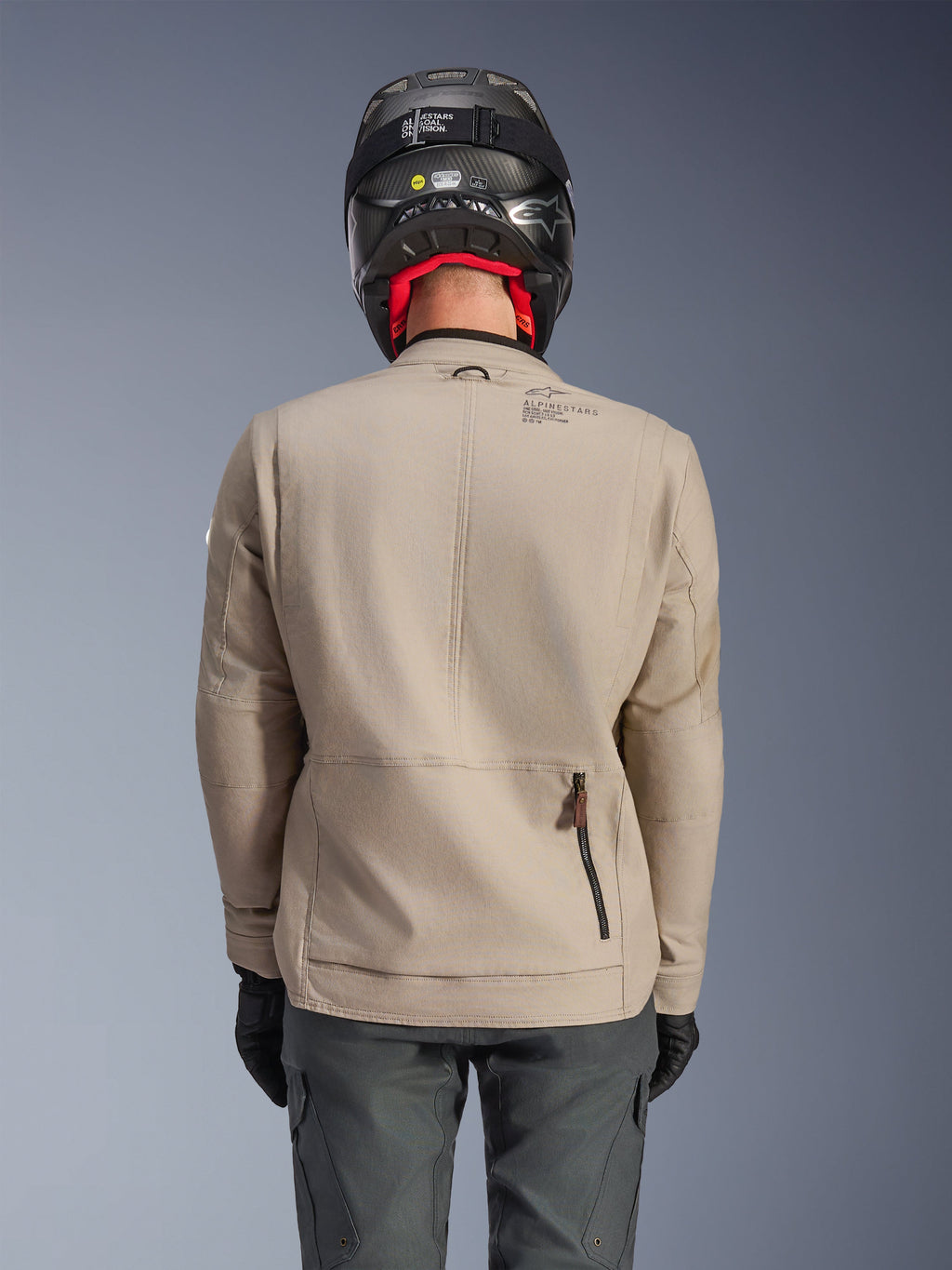 Alpinestars Borrego Drystar®-jacka, äventyrsmotorcykeljacka, Warm Gray, ryggvy som visar dragkedja för bakre fack och Alpinestars-logotyp, bärs med en svart hjälm och grafitgrå körbyxor