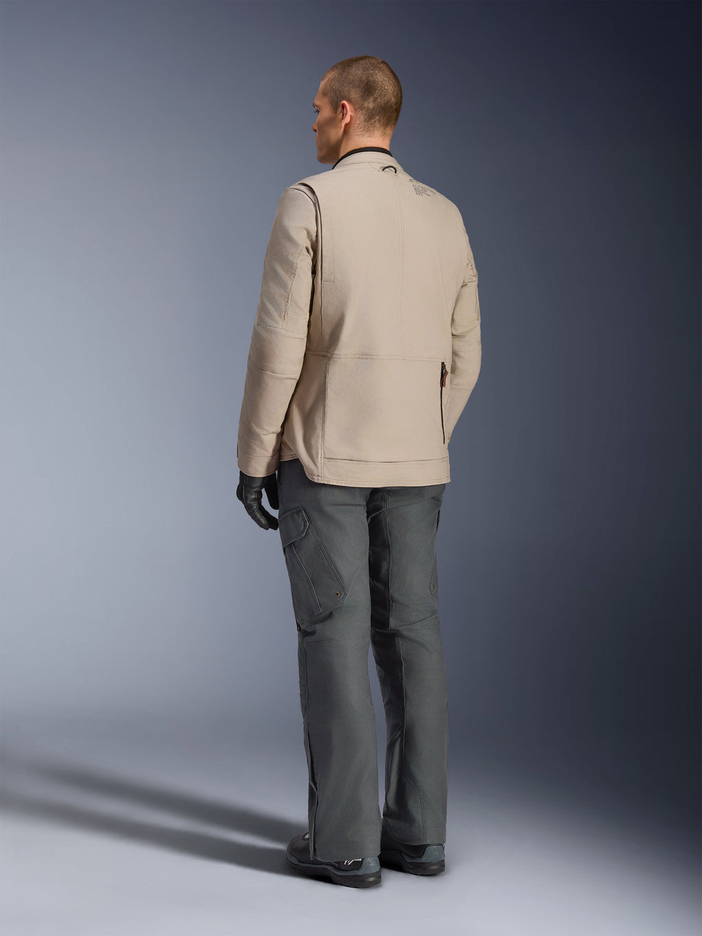 Alpinestars Borrego Drystar®-jacka i färgen Warm Gray, vy bakifrån som visar den robusta vaxade canvas-strukturen, expansionspaneler på axlarnas baksida och en stor bakficka med dragkedja, bärs av en modell med koksgrå äventyrsbyxor och svarta handskar.