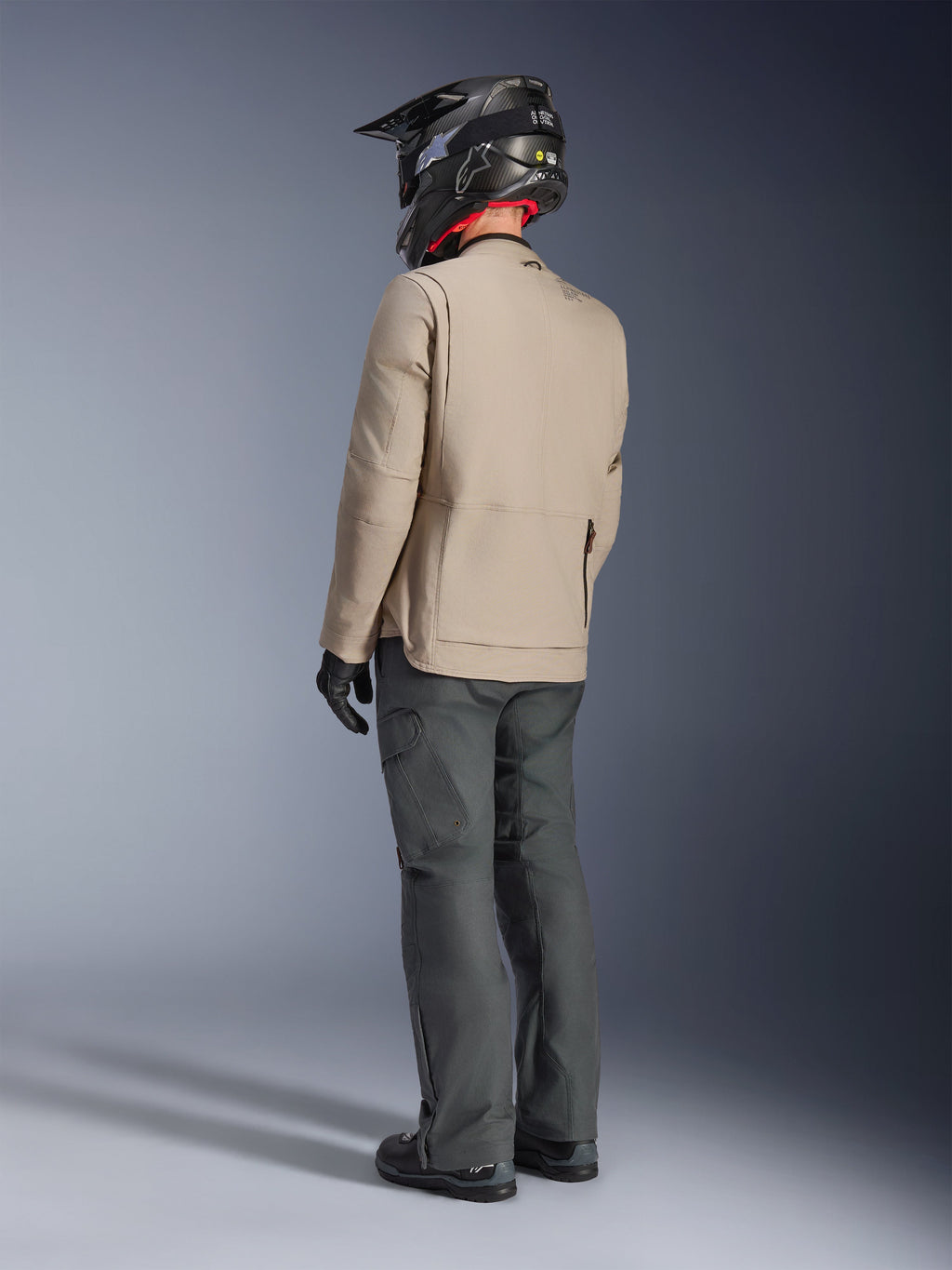 Alpinestars Borrego Drystar®-jacka, Warm Gray äventyrsmotorcykeljacka, ryggvy av förare med mörkgrå byxor och en svart hjälm, med en robust vaxad canvas-look och funktionell design.