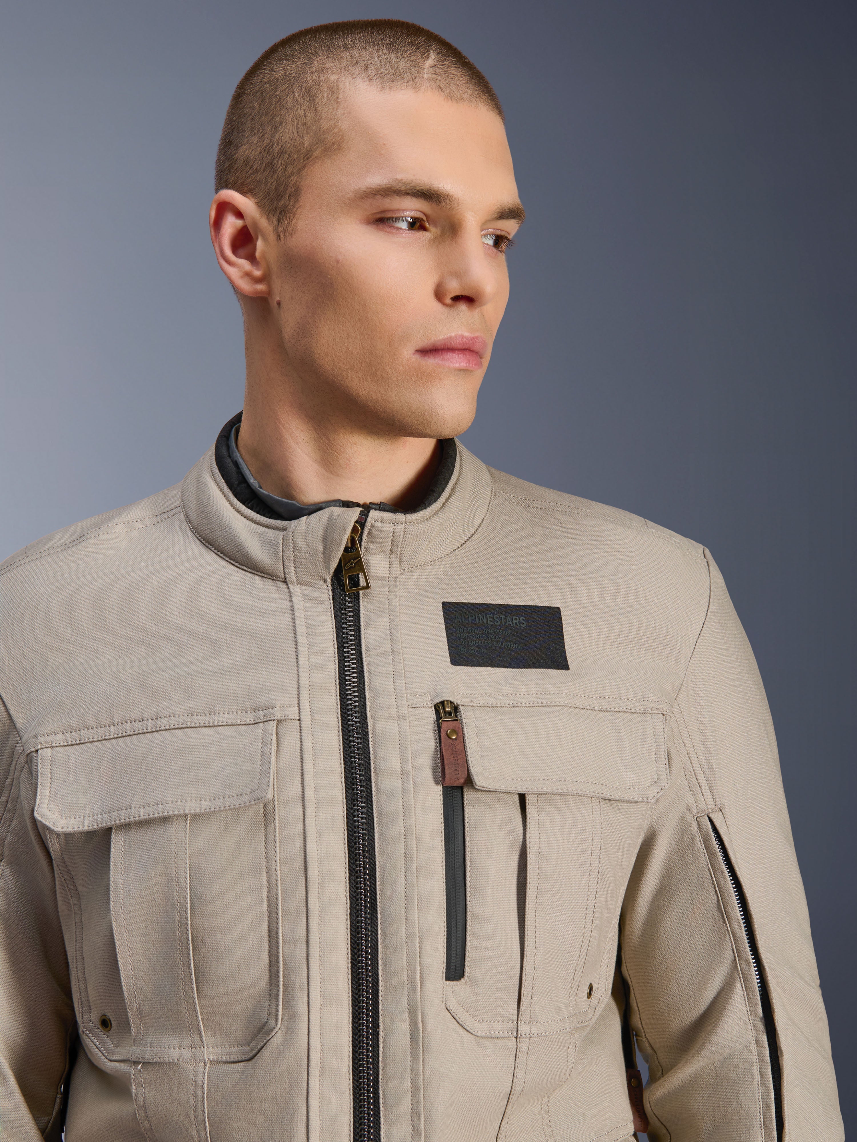 Manlig modell som bär Alpinestars Borrego Drystar®-jacka i Warm Gray, äventyrsmotorcykeljacka med en klassisk vaxad canvas-look, funktionella cargofickor på bröstet och ett svart Alpinestars-logomärke.