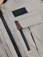 Alpinestars Borrego Drystar® Jacka, Warm Gray, närbild av bröstpartiet med en svart Alpinestars-märkespatch, bröstficka med lock och en vertikal dragkedja med en dragtapp i brunt läder, hållbar konstruktion i grå textilcanvas.