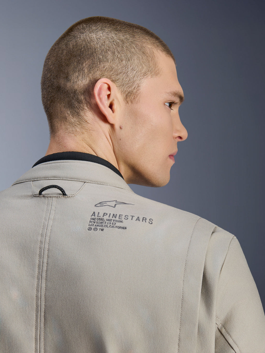 Alpinestars Borrego Drystar®-jacka i Warm Gray, närbild på axelpartiet bak, texturerat vaxat canvasmaterial, med svart Alpinestars-logotyp och heritagetext, bärs av modell, motorcykeljacka för äventyr