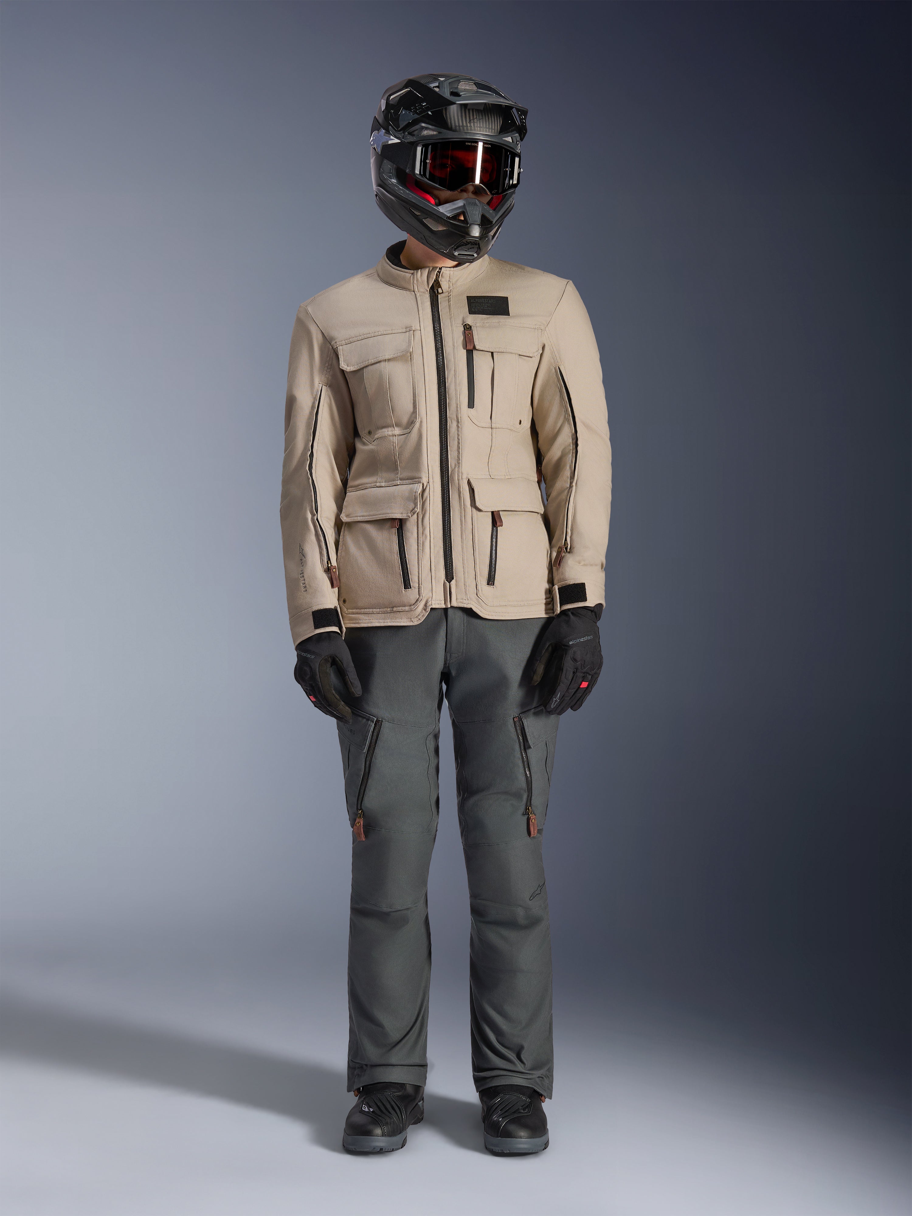 Alpinestars Borrego Drystar®-jacka i Warm Gray, äventyrsmotorcykeljacka med fyra cargo-fickor framtill, kombinerad med mörkgrå mc-byxor, svart offroad-hjälm och svarta handskar, med en robust vaxad canvas-look