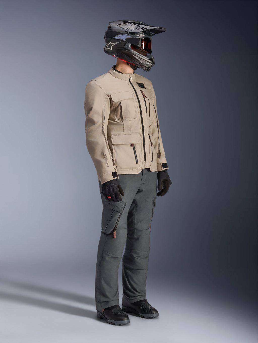 Motorcyklist klädd i Alpinestars Borrego Drystar®-jacka i Warm Gray, adventure-motorcykeljacka med vaxad canvas-textur och praktiska fickor, stylad med mörkgrå mc-byxor, kolfiberhjälm, svarta handskar och svarta stövlar, helkroppsbild i studiomiljö av en komplett outfit för äventyrstouring.