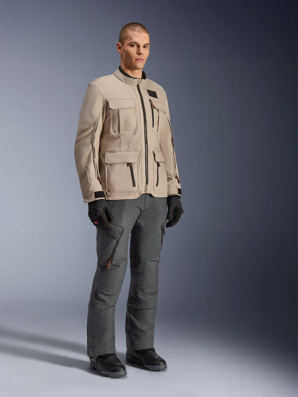 Alpinestars Borrego Drystar®-jacka i Warm Gray, äventyrsmotorcykeljacka med vaxad canvas-look och flera praktiska fickor, buren av en modell med mörkgrå körbyxor, svarta handskar och svarta stövlar, studioporträtt i helfigur