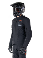Alpinestars AMT-8 Stretch Drystar® XF Jacka, svart, bärs av en förare med matchande äventyrsbyxor och handskar, svart motocrosshjälm och goggles, ventilationsöppningar med dragkedja på bröstet och armarna, Alpinestars Motion Technology-branding med en orange logotyp, premium adventure-touringutrustning