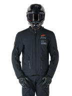 Alpinestars AMT-8 Stretch Drystar® XF-jacka, svart motorcykeljacka med orange Alpinestars-logotyp och grå AMT-branding, hög krage med dragkedja fram och ventilationsöppningar, bärs med svart hjälm och handskar, skyddande utrustning för adventure touring