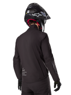 Alpinestars AMT-8 Stretch Drystar® XF-foder, svart vattentätt jackfoder, vy bakifrån på en förare med svart hjälm och handskar, Alpinestars Motion Technology-märkning på nedre delen av ryggen, minimalistisk och funktionell design för äventyrskörning.