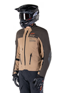 Alpinestars AMT-8 Stretch Drystar® XF jacka, äventyrsmotorcykeljacka, mörkbrun och mörk khaki, bärs med en svart terränghjälm och goggles, med Alpinestars Motion Technology-logotyp och ventilationsöppningar med dragkedja.