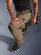 Alpinestars AMT-8 Stretch Drystar® XF-byxor i Walnut Black, med Alpinestars Motion Technology (AMT)-logotyp på låret, långa ventilationsöppningar med dragkedja och slitstarka knäpaneler i läder, visas på en förare i knästående position för att framhäva flexibilitet.