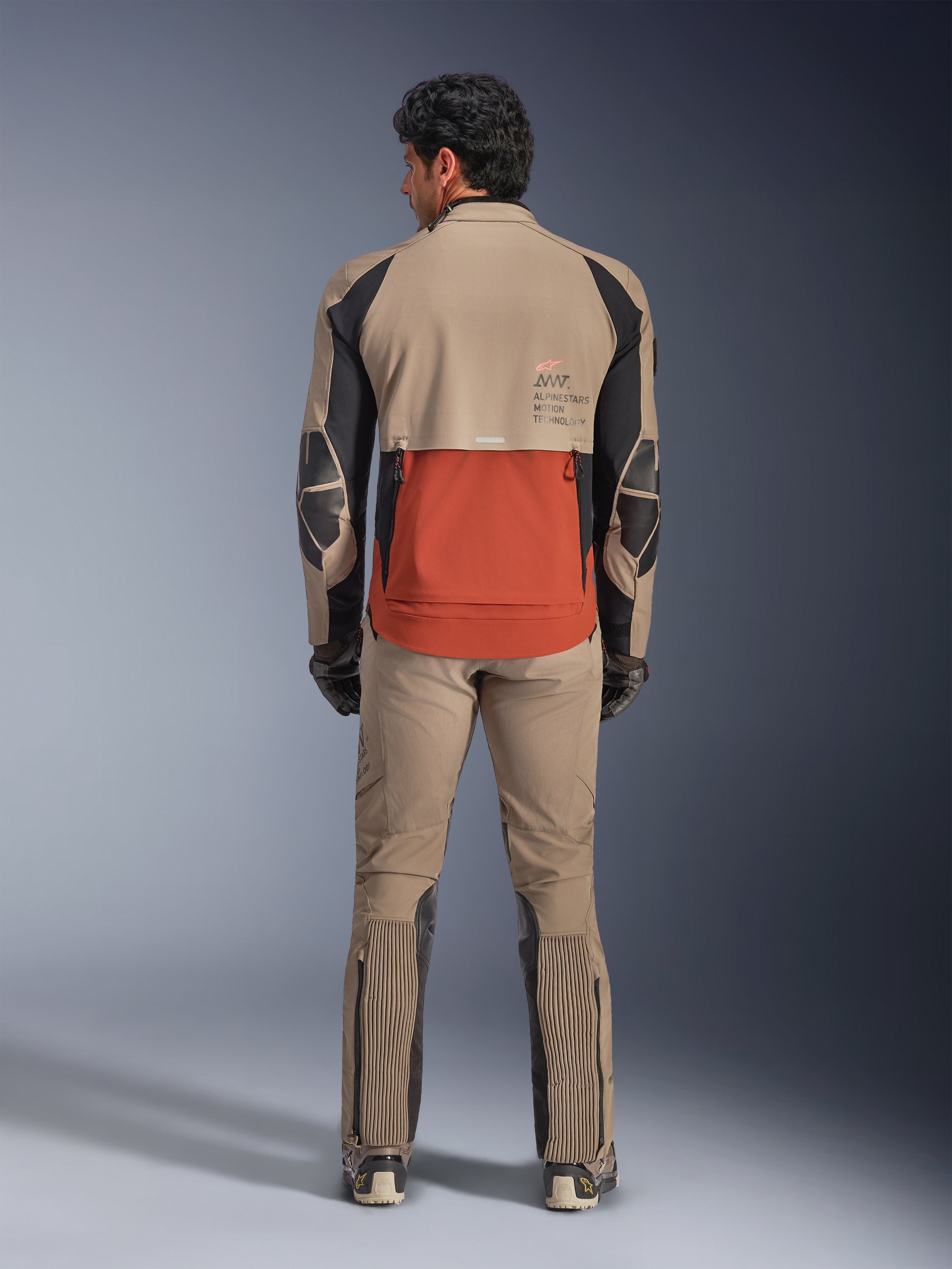 Alpinestars AMT-8 Stretch Drystar® XF-jacka, Walnut Rust Black, ryggvy på en förare med matchande adventure-byxor och handskar, med en framträdande rostfärgad bakpanel, valnötsfärgade övre sektioner och svarta stretchdetaljer, designad för långdistans touring.