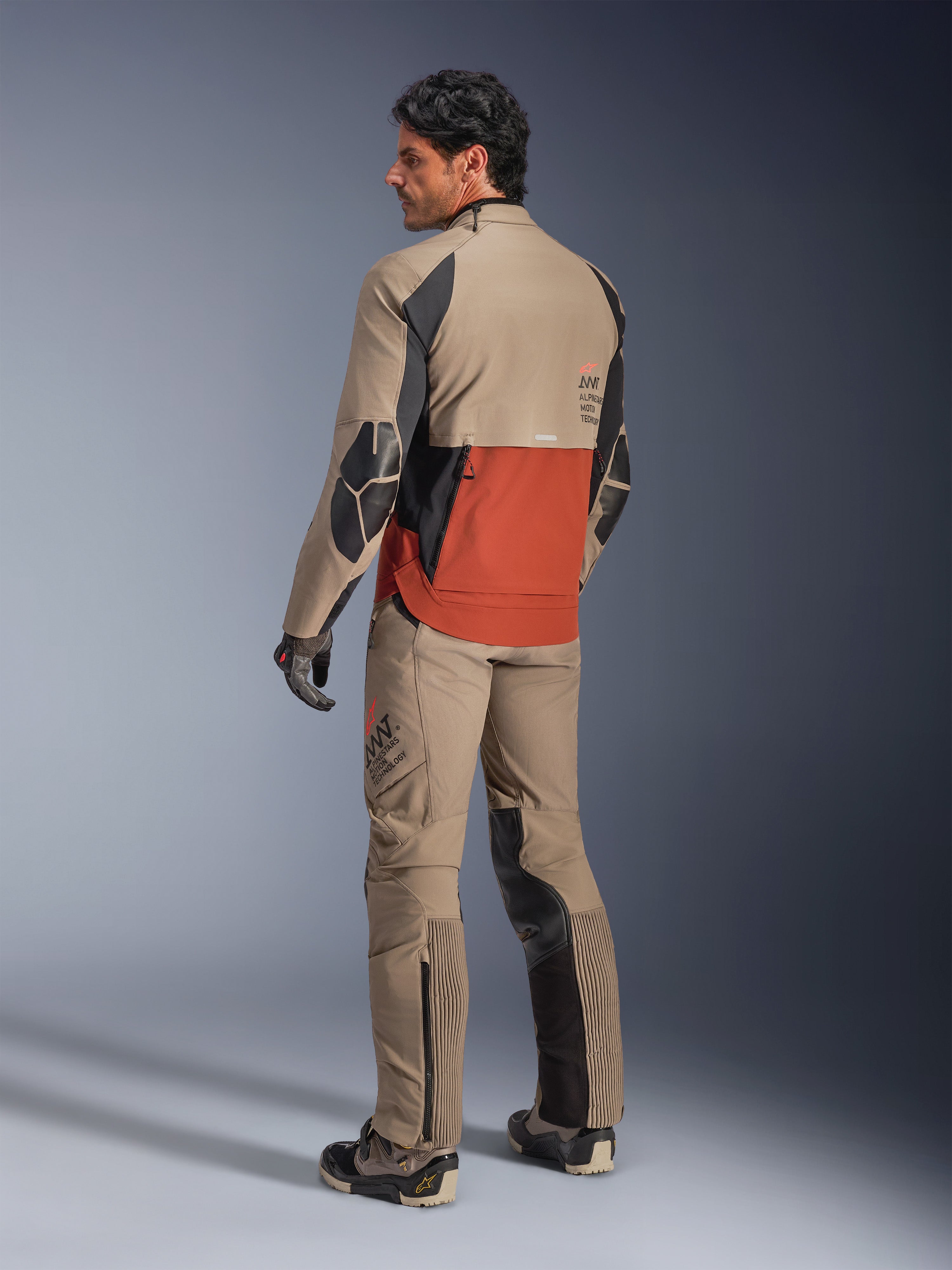 Alpinestars AMT-8 Stretch Drystar® XF-jacka, äventyrsmotorcykeljacka, valnöt, rost och svart, baksida på modell som bär matchande äventyrsbyxor och stövlar, textilutrustning med hög andningsförmåga för touring
