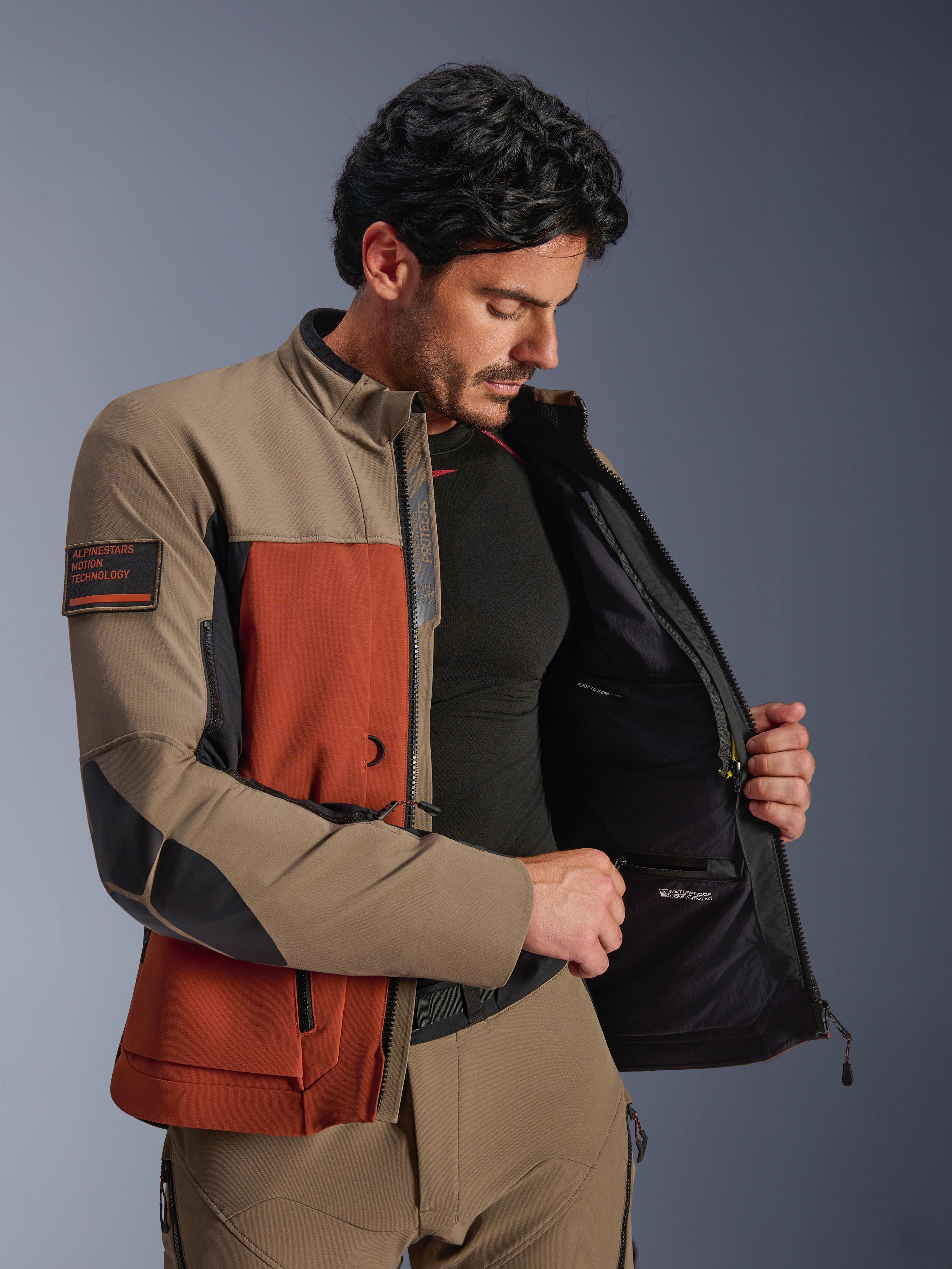 Alpinestars AMT-8 Stretch Drystar® XF-jacka, Walnut Rust Black, man som bär jackan öppen för att visa det inre vattentäta facket och det tekniska fodret, med 'Alpinestars Motion Technology'-märke på ärmen, äventyrsmotorcykelutrustning