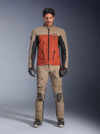Alpinestars AMT-8 Stretch Drystar® XF Jacka, Walnut Rust Black äventyrsjacka för motorcykel, bärs av modell med matchande tekniska byxor och handskar, med en flerfärgad stretch-textildesign, främre cargofickor och logotyper, vy framifrån.