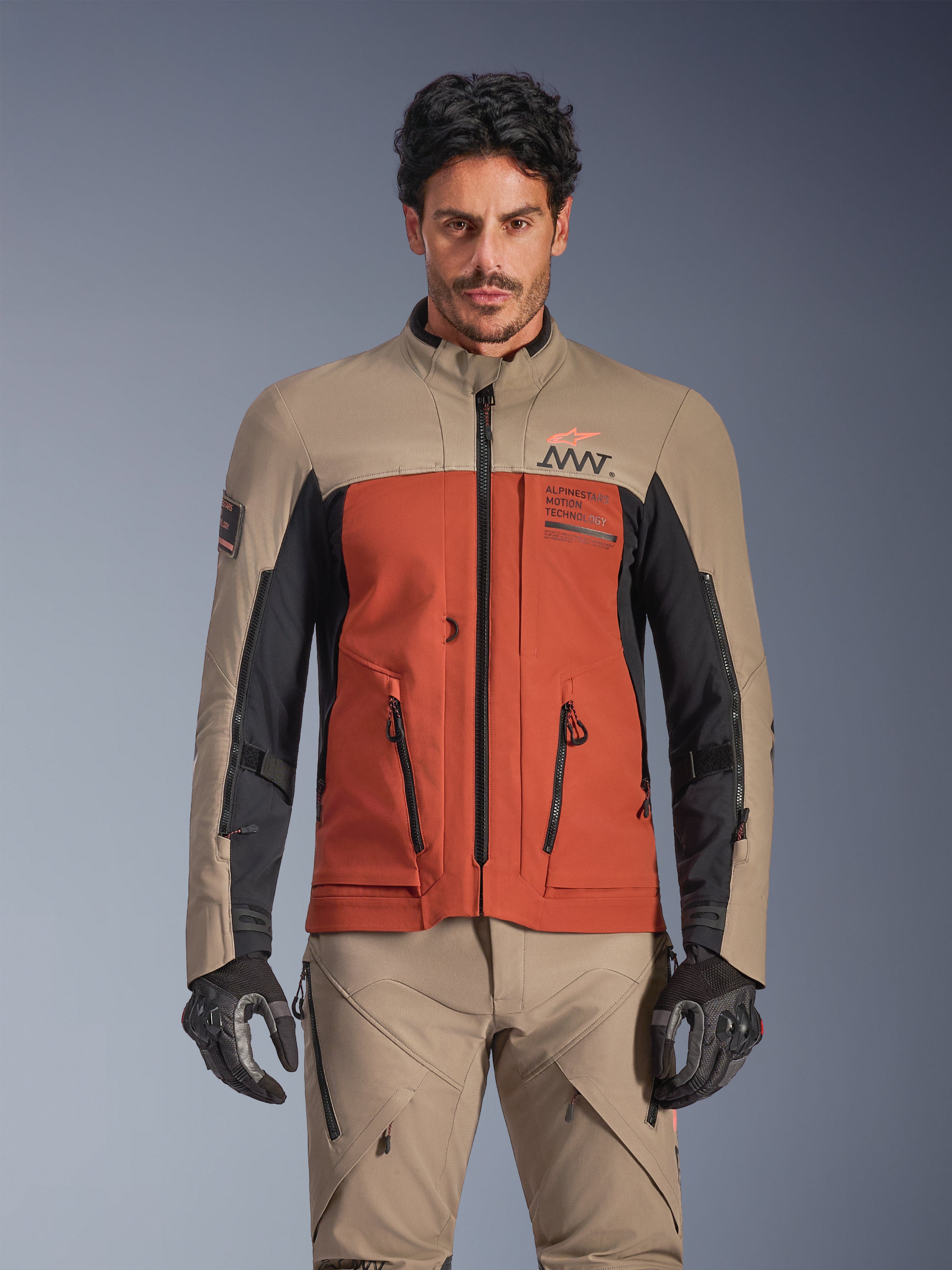 Alpinestars AMT-8 Stretch Drystar® XF-jacka, Walnut Rust Black adventure touring-jacka, bärs av en manlig modell med matchande byxor och handskar, trefärgad design i beige, rostbrunt och svart med AMT-logotyp på bröstet, aktiv touring-passform för motorcykelkörning