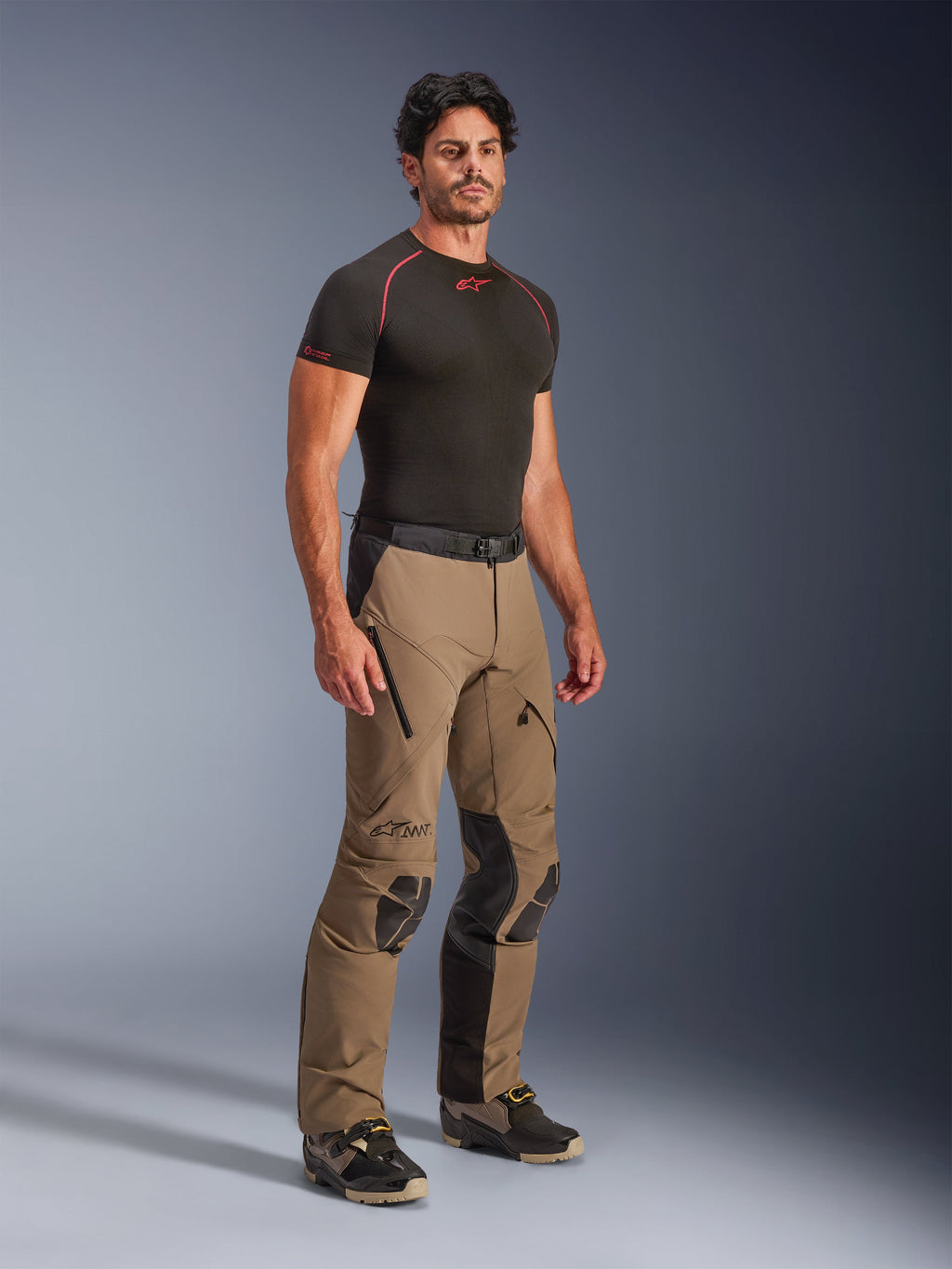 Alpinestars AMT-8 Stretch Drystar® XF-byxor i Walnut Black, adventure touring-motorcykelbyxor som bärs av en modell med en svart underställströja, med stretchmaterial, dragkedjeförsedda ventilationsöppningar på låren och skyddande knäpaneler.