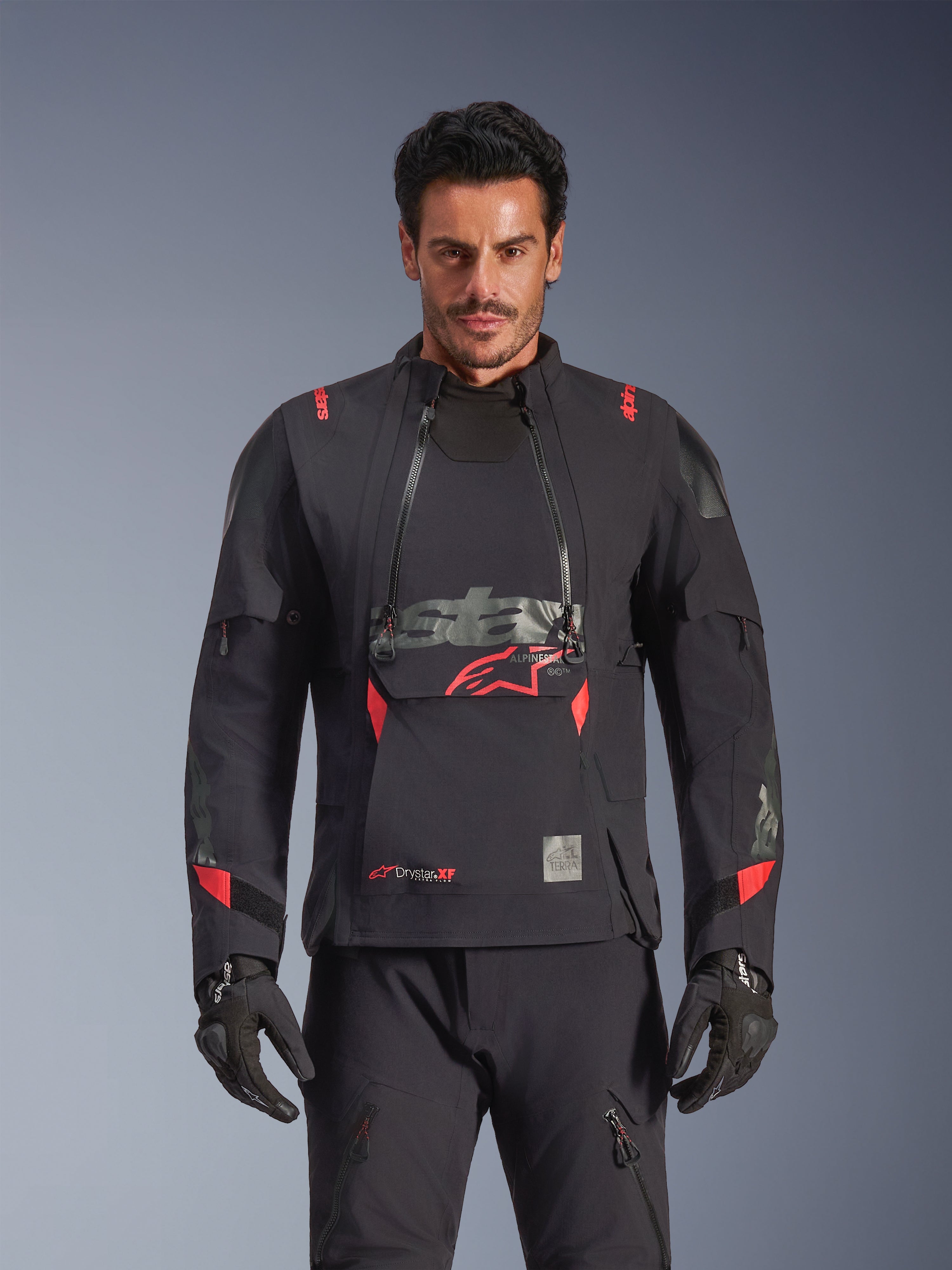 Alpinestars Halo Pro Drystar® XF laminerad jacka, äventyrsmotorcykeljacka, Black Dark Shadow med röda detaljer, manlig modell i komplett teknisk utrustning med matchande handskar och byxor, frontvy som framhäver modulära ventilationspaneler på bröstet och vattentät konstruktion.