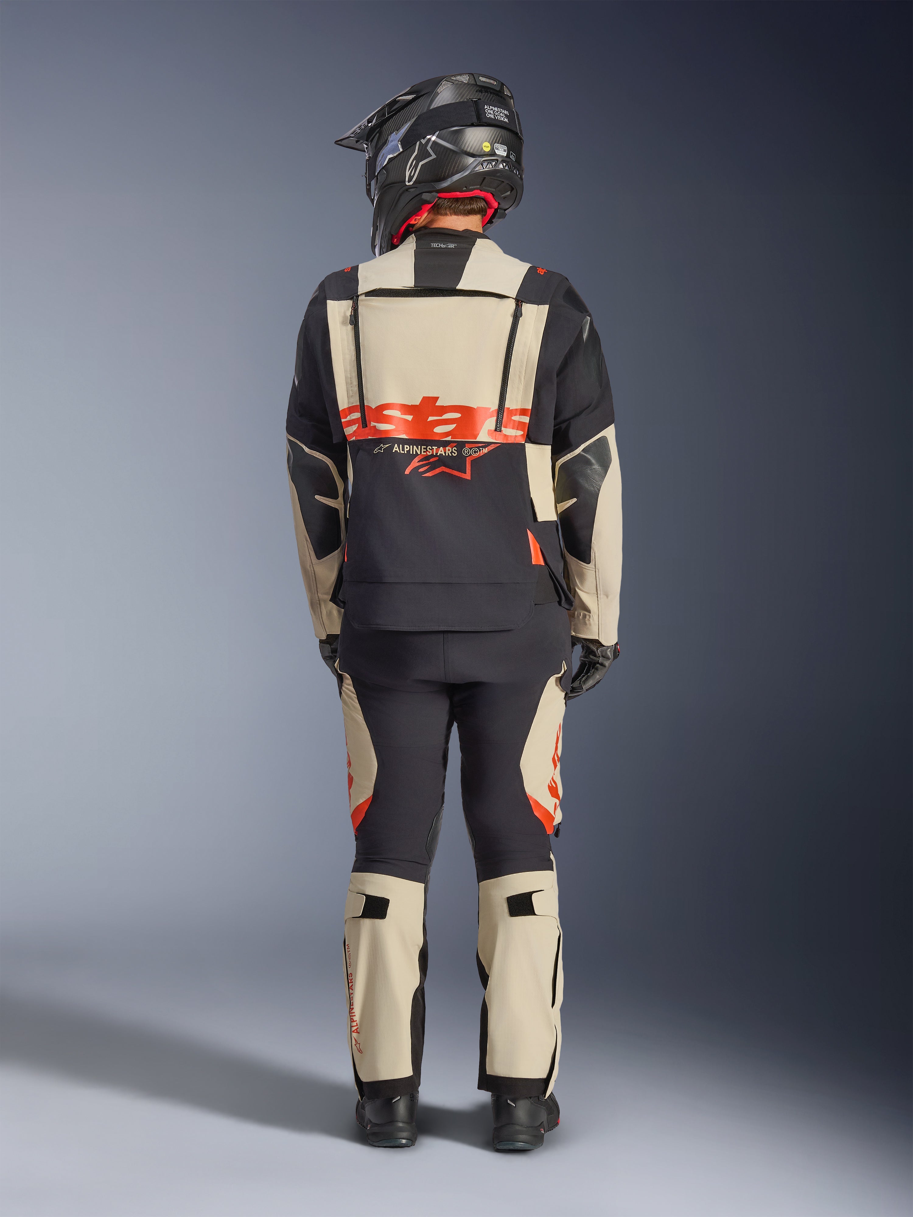 Alpinestars Halo Pro Drystar® XF laminerad jacka, äventyrsmotorcykeljacka, Mastic Black, ryggvy av en förare i matchande byxor och svart hjälm, med framträdande orange Alpinestars-logotyp och modulära ventilationspaneler för mångsidig äventyrskörning.