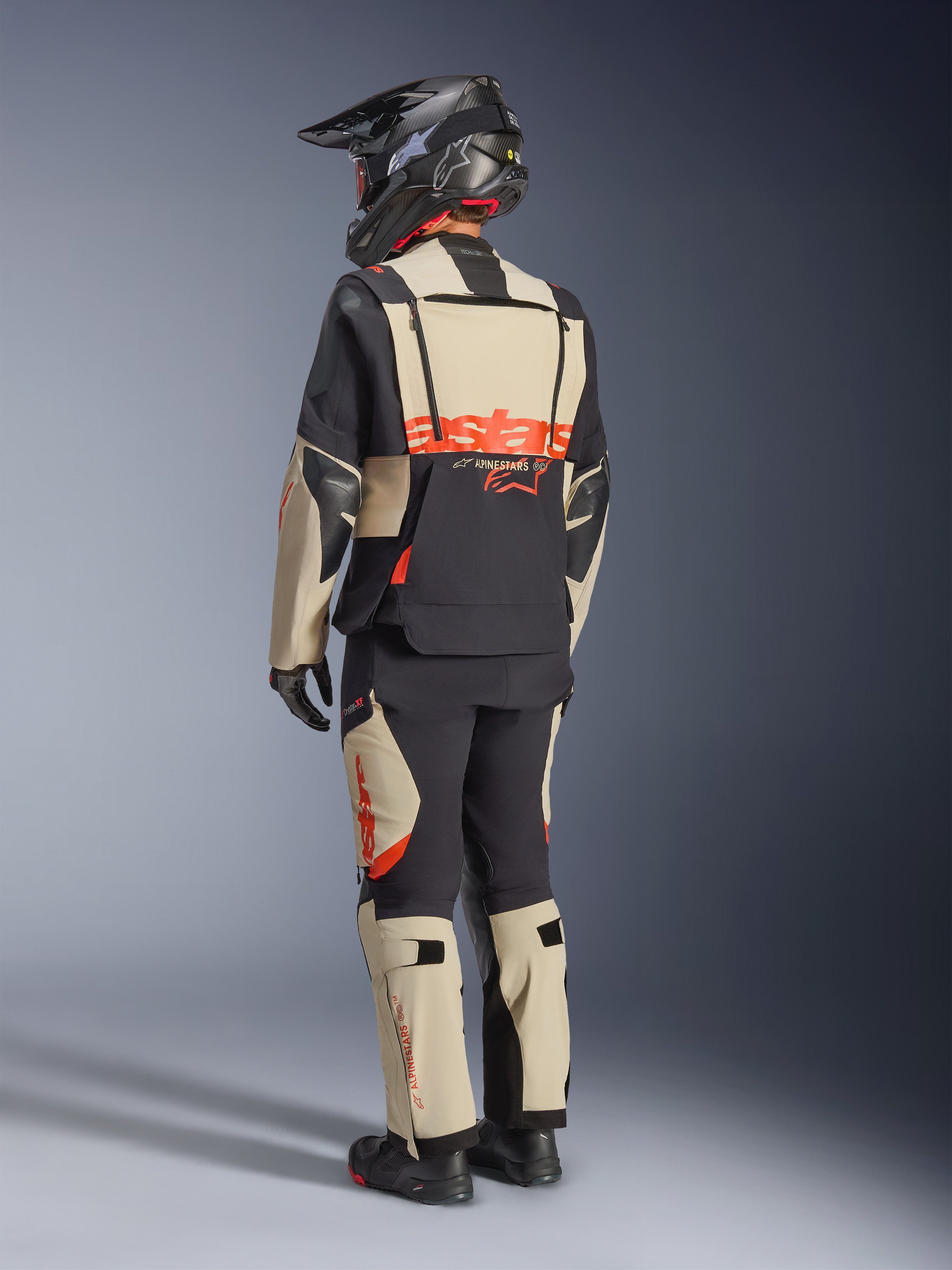 Alpinestars Halo Pro Drystar® XF laminerad jacka, Mastic Black, vy bakifrån av en förare iklädd ett komplett äventyrsställ med matchande byxor, svart äventyrshjälm och stövlar, med en framträdande röd Alpinestars-logotyp över ryggen.
