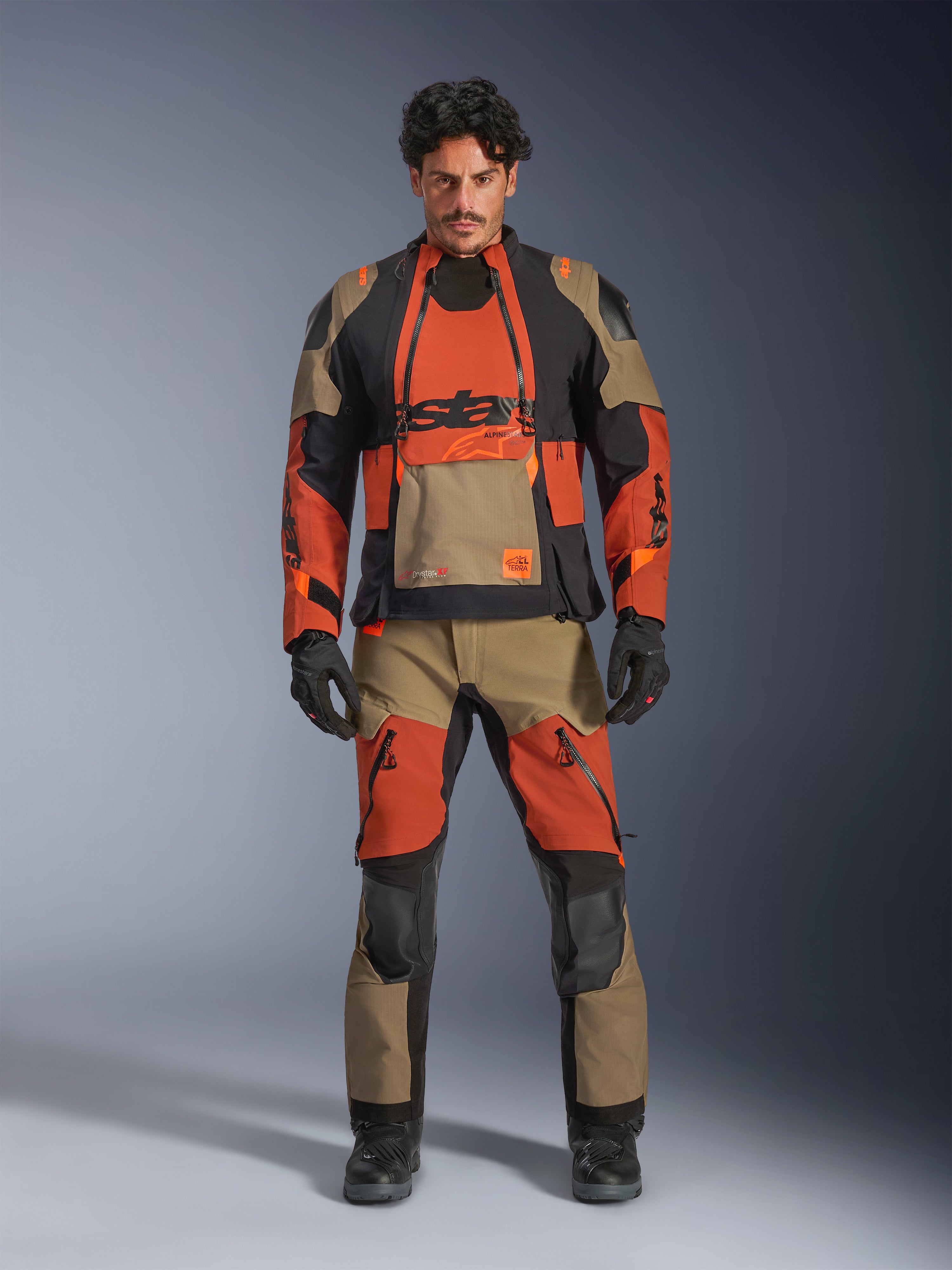 Manlig modell bär Alpinestars Halo Pro Drystar® XF-laminerad jacka och matchande byxor i Walnut Rust Black, modulär äventyrsmotorcykelutrustning med främre ventilationspaneler och dragkedjor, stylad med svarta motorcykelhandskar och stövlar.