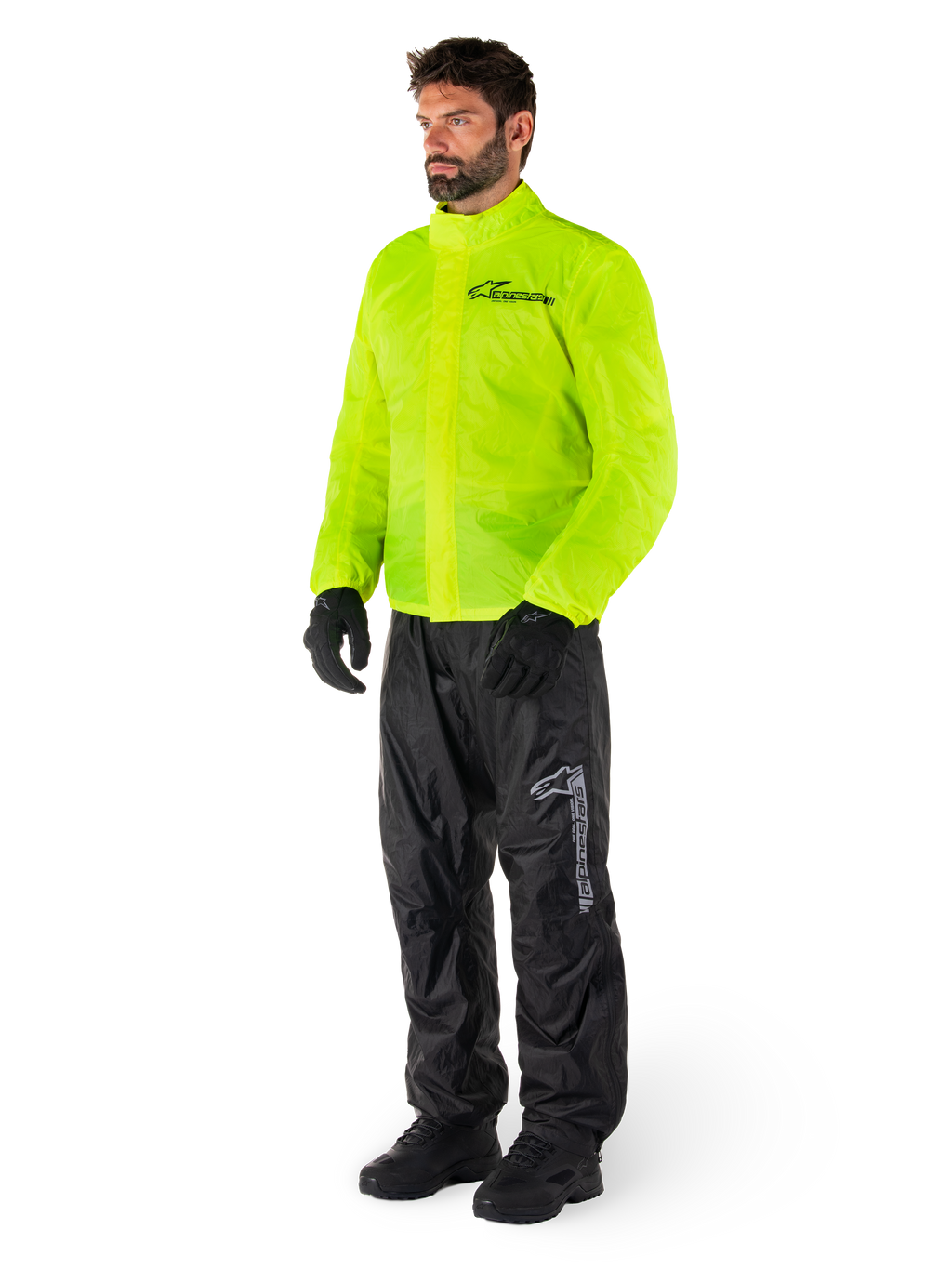 Man i Alpinestars Hurricane Rain V2-jacka i Yellow Fluo och Hurricane Rain V2-byxor i svart, vattentät motorcykelregnutrustning med hög synlighet, med reflekterande logotyper på bröst och lår, komplett outfit med svarta handskar och stövlar.