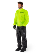 Man i Alpinestars Hurricane Rain V2-jacka i Yellow Fluo och Hurricane Rain V2-byxor i svart, vattentät motorcykelregnutrustning med hög synlighet, med reflekterande logotyper på bröst och lår, komplett outfit med svarta handskar och stövlar.