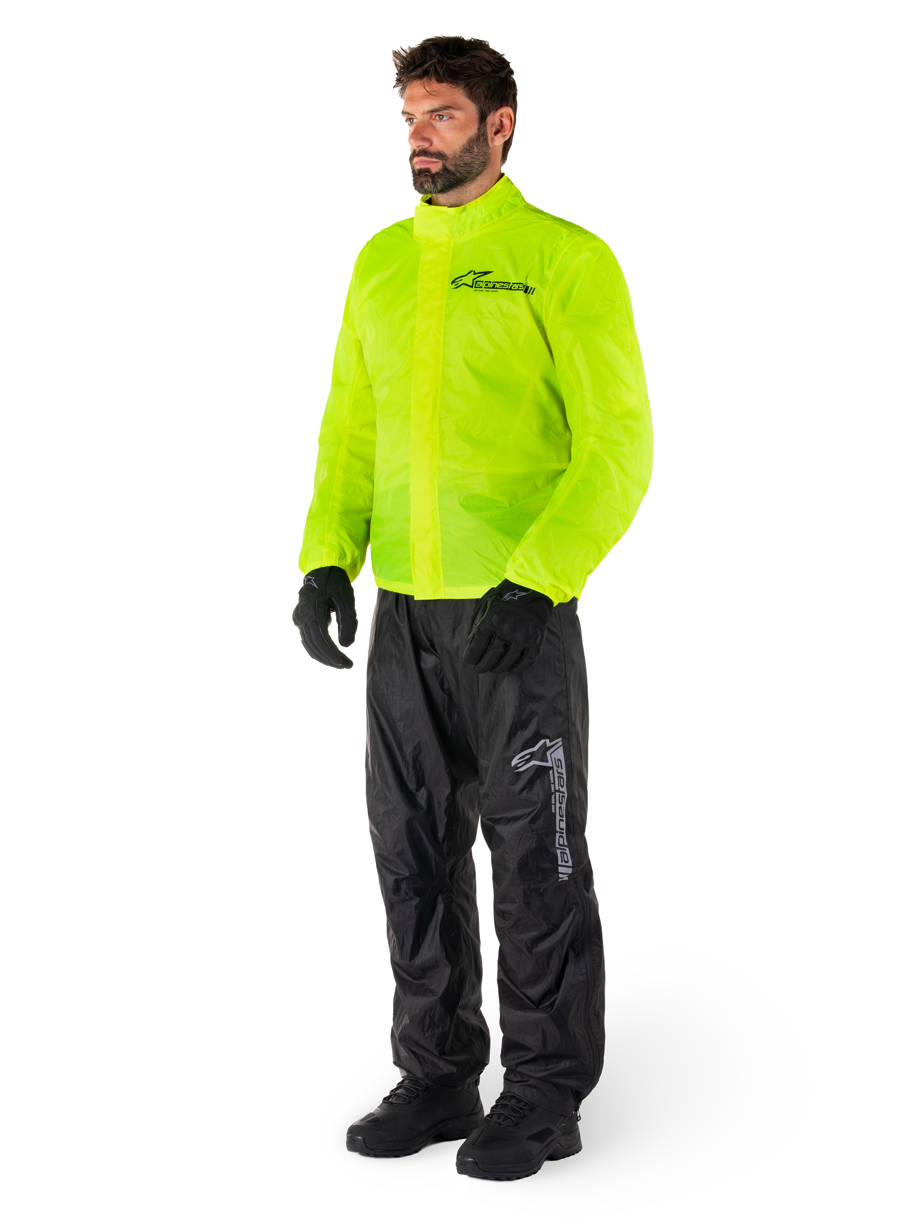 Man i Alpinestars Hurricane Rain V2-jacka i Yellow Fluo och Hurricane Rain V2-byxor i svart, vattentät motorcykelregnutrustning med hög synlighet, med reflekterande logotyper på bröst och lår, komplett outfit med svarta handskar och stövlar.
