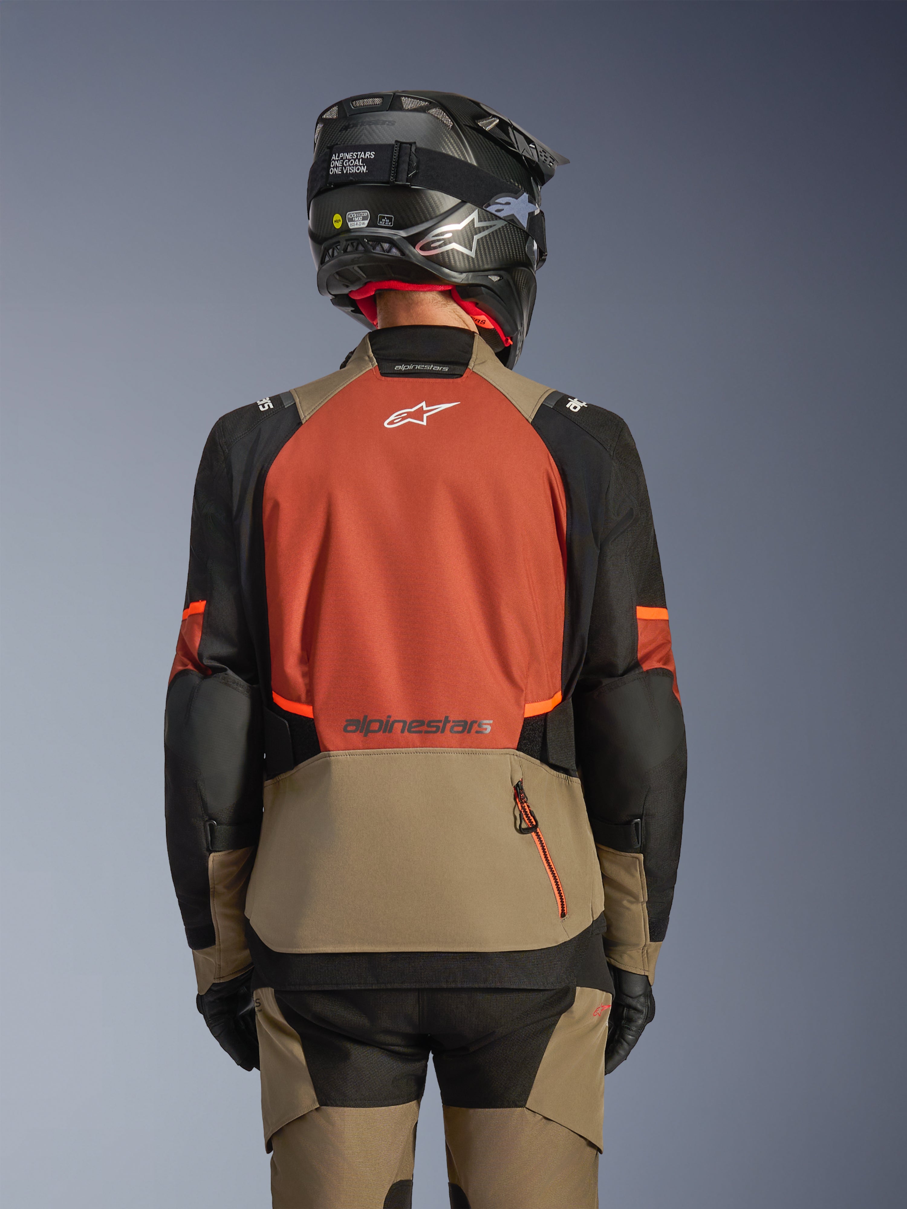 Baksida av Alpinestars Andes V4 Drystar®-jacka i Walnut Rust Black, adventure touring-jacka med en stor rostfärgad ryggpanel och vit logotyp, valnötsfärgad nedre del med en vertikal dragkedjeficka, svarta ärmar med orangea detaljer, stylad med matchande byxor och en svart hjälm för körning i alla väder.