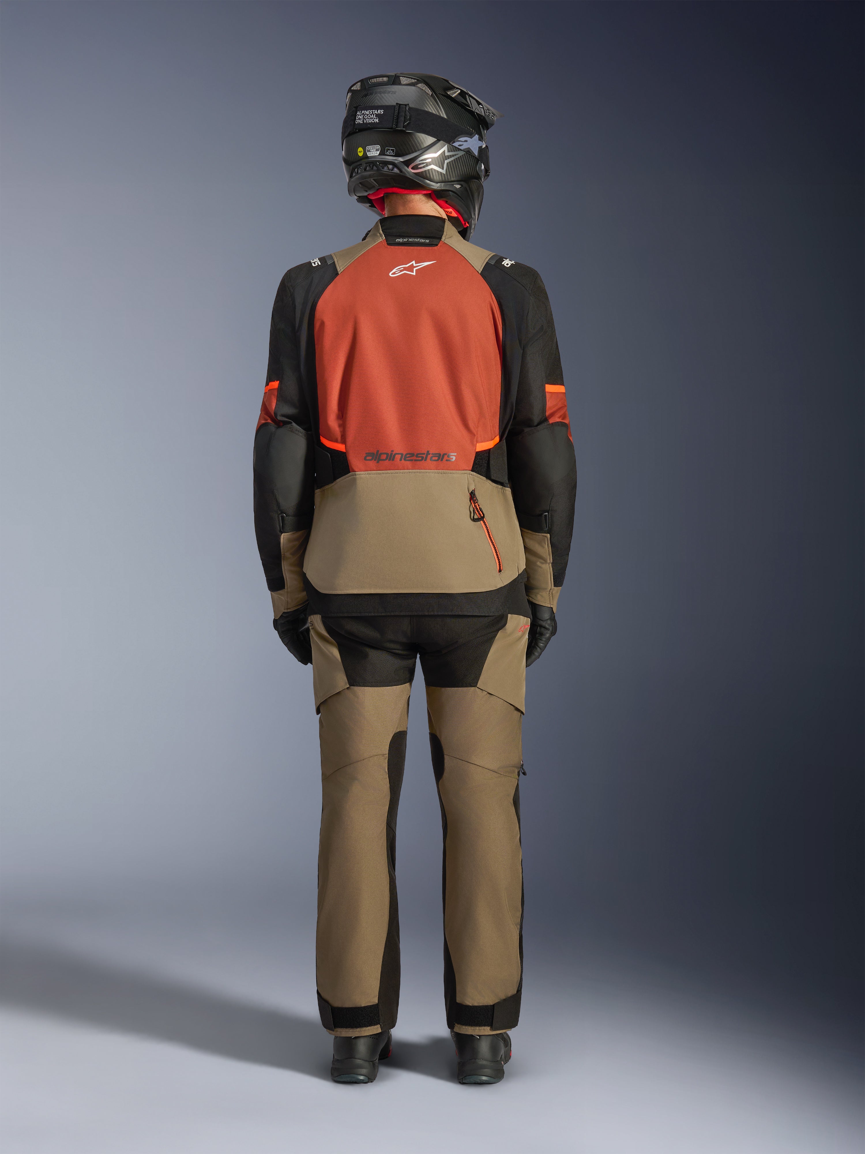 Bakifrån-vy av en förare som bär Alpinestars Andes V4 Drystar® Jacket i Walnut Rust Black, tillsammans med matchande valnötsfärgade äventyrsbyxor och en svart hjälm, vilket visar jackans rostfärgade ryggpanel, stora bakre förvaringsficka och reflekterande logotyper.