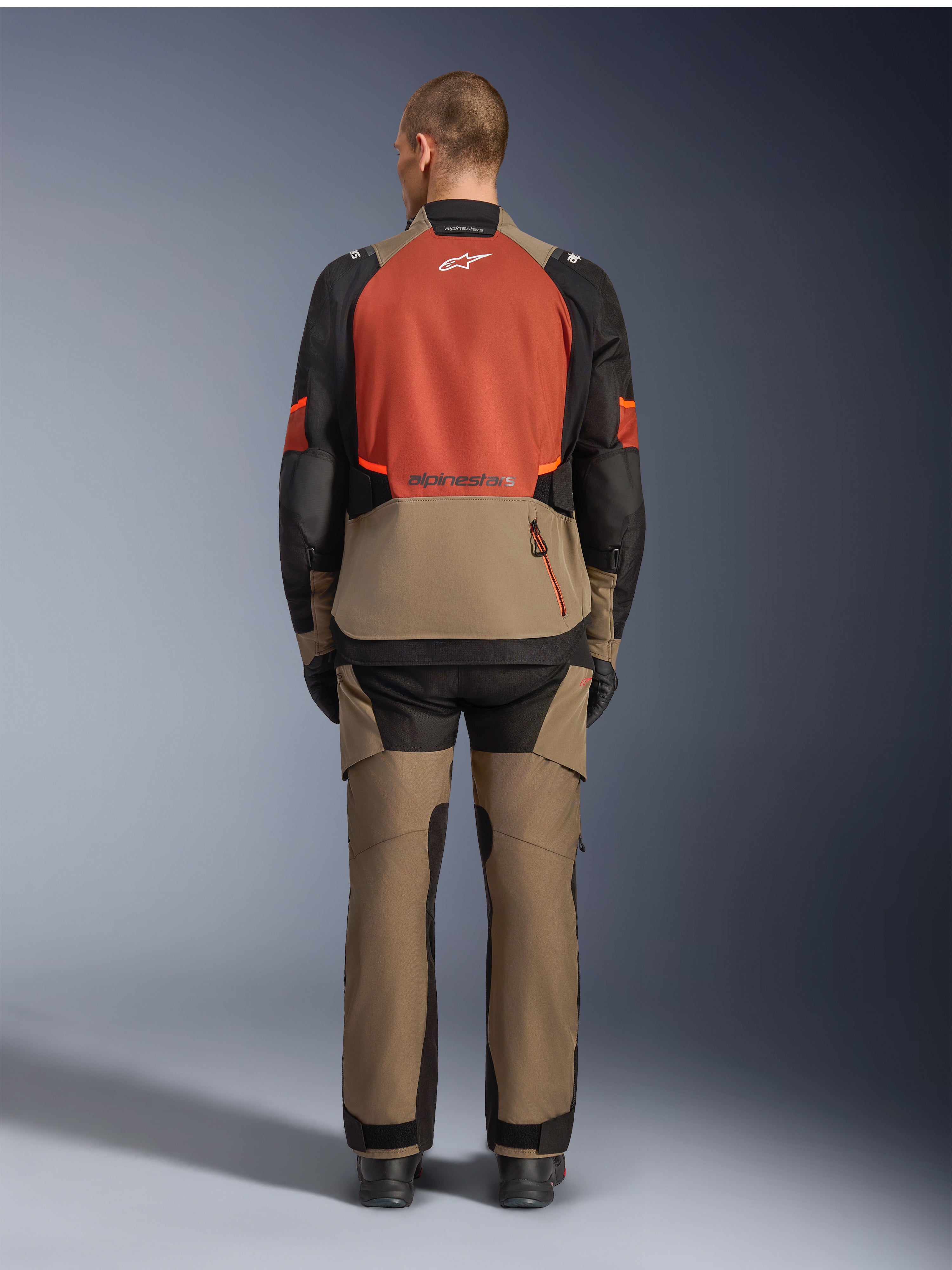 Alpinestars Andes V4 Drystar®-jacka, Walnut Rust Black, baksida av motorcykeljacka för touring med rostfärgad övre ryggpanel och valnötsfärgad nedre sektion, bärs med matchande äventyrsbyxor och svarta stövlar.