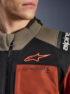 Alpinestars Andes V4 Drystar®-jacka i Walnut Rust Black, närbild på övre bröst och axel, med orange A-star-logotyp på en svart panel och Alpinestars-märkning på en rostfärgad del, detalj av äventyrsmotorcykelutrustning