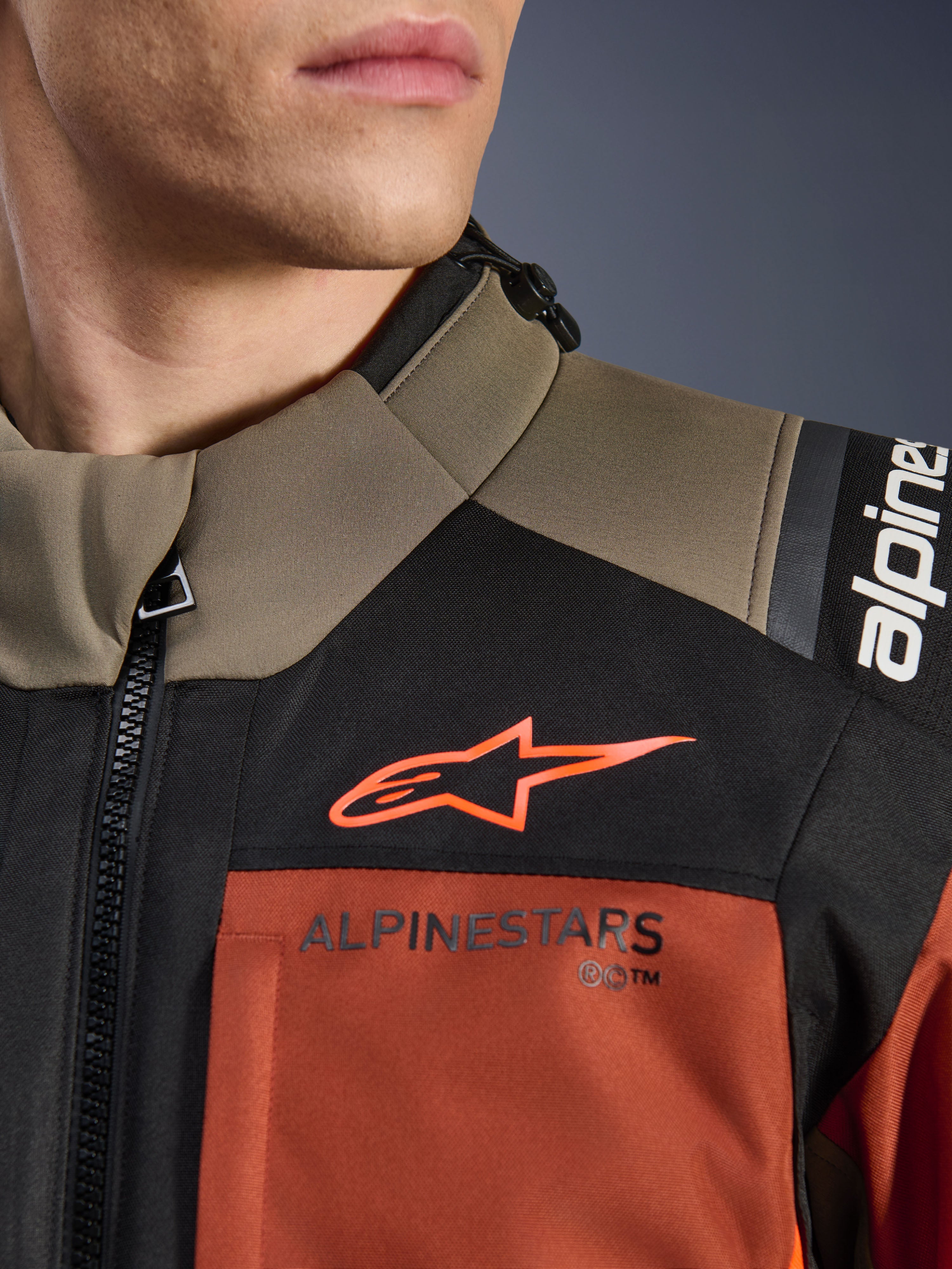 Alpinestars Andes V4 Drystar®-jacka i Walnut Rust Black, närbild på övre bröst och axel, med orange A-star-logotyp på en svart panel och Alpinestars-märkning på en rostfärgad del, detalj av äventyrsmotorcykelutrustning